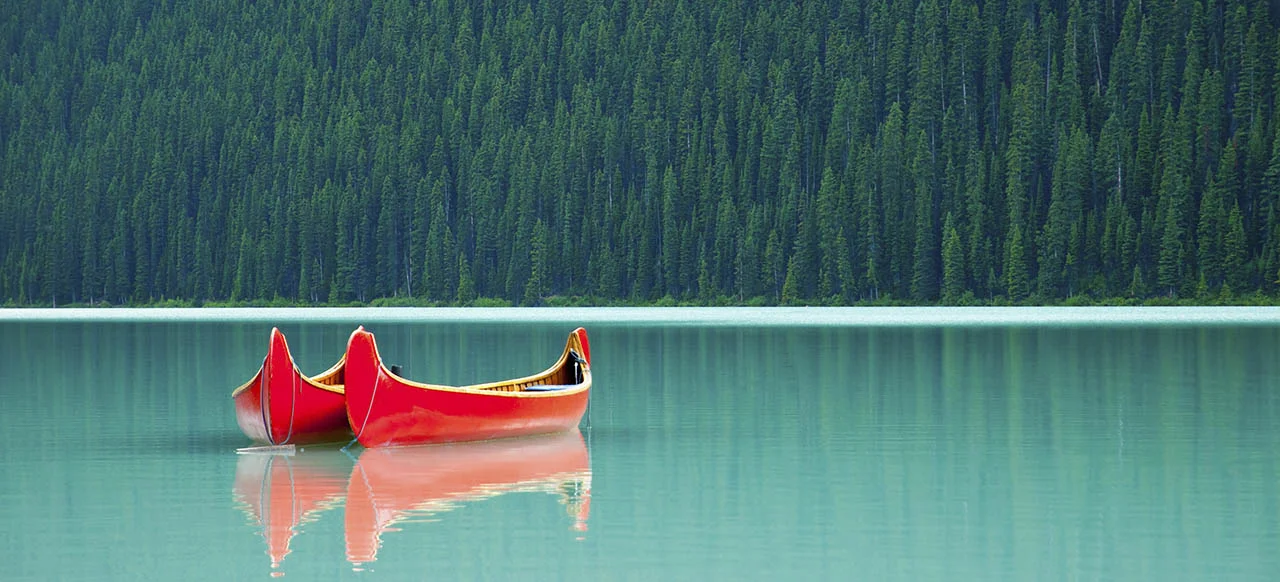 Image of two canoes sitting next to each other on a calm lake with trees in the background