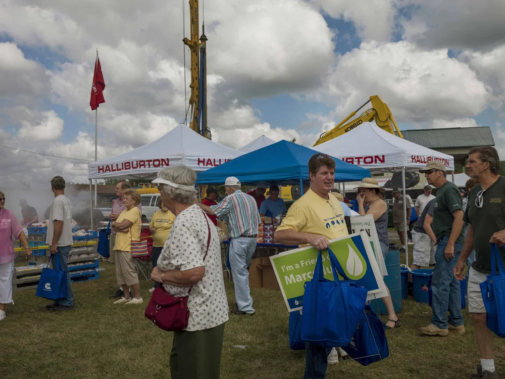 An EOG Cabot Oil and Gas Community picnic at the Hartford Fairgrounds attracts shale gas businesses and residents. Here picnic goers, one carrying a sign, pass in front of the Halliburton barbecue pit. Around 7,000 people attended. Hartford, Susquehanna County, 2012. © Nina Berman/MSDP 2012