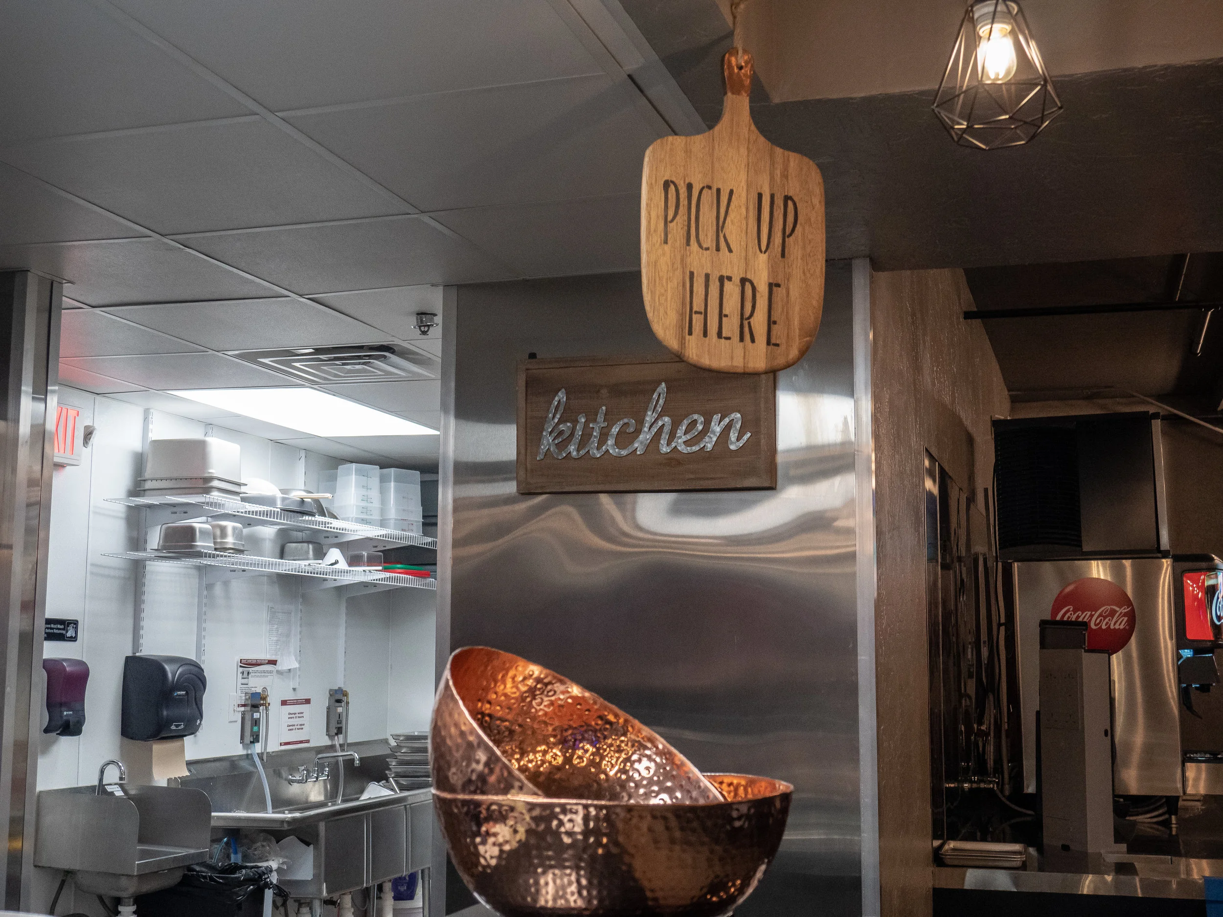 A sign hanging from the ceiling that says “pick up here” and a sign on a wall behind that saying “kitchen.”