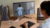 A person working on a computer with a large monitor displaying 3D character modeling and animation in a digital art program. The desk has a glass of water, a smartphone, and a notebook.