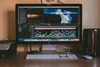 A computer monitor displaying video editing software with a timeline, video clips, and color grading tools. A keyboard, mouse, and external speakers are on the desk.