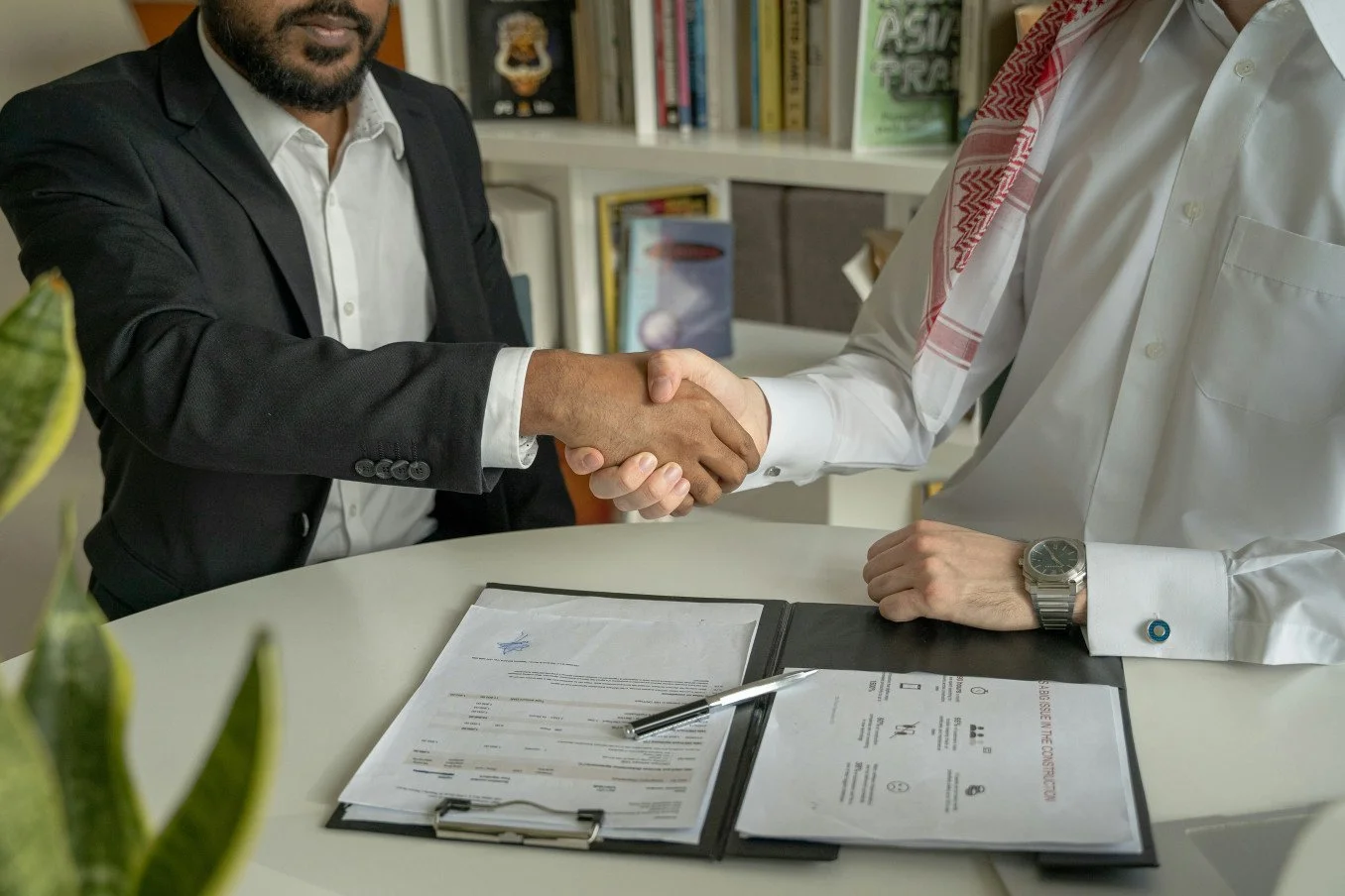 Two men in business attire shaking hands across a table with documents, pens, and a bookshelf in the background.