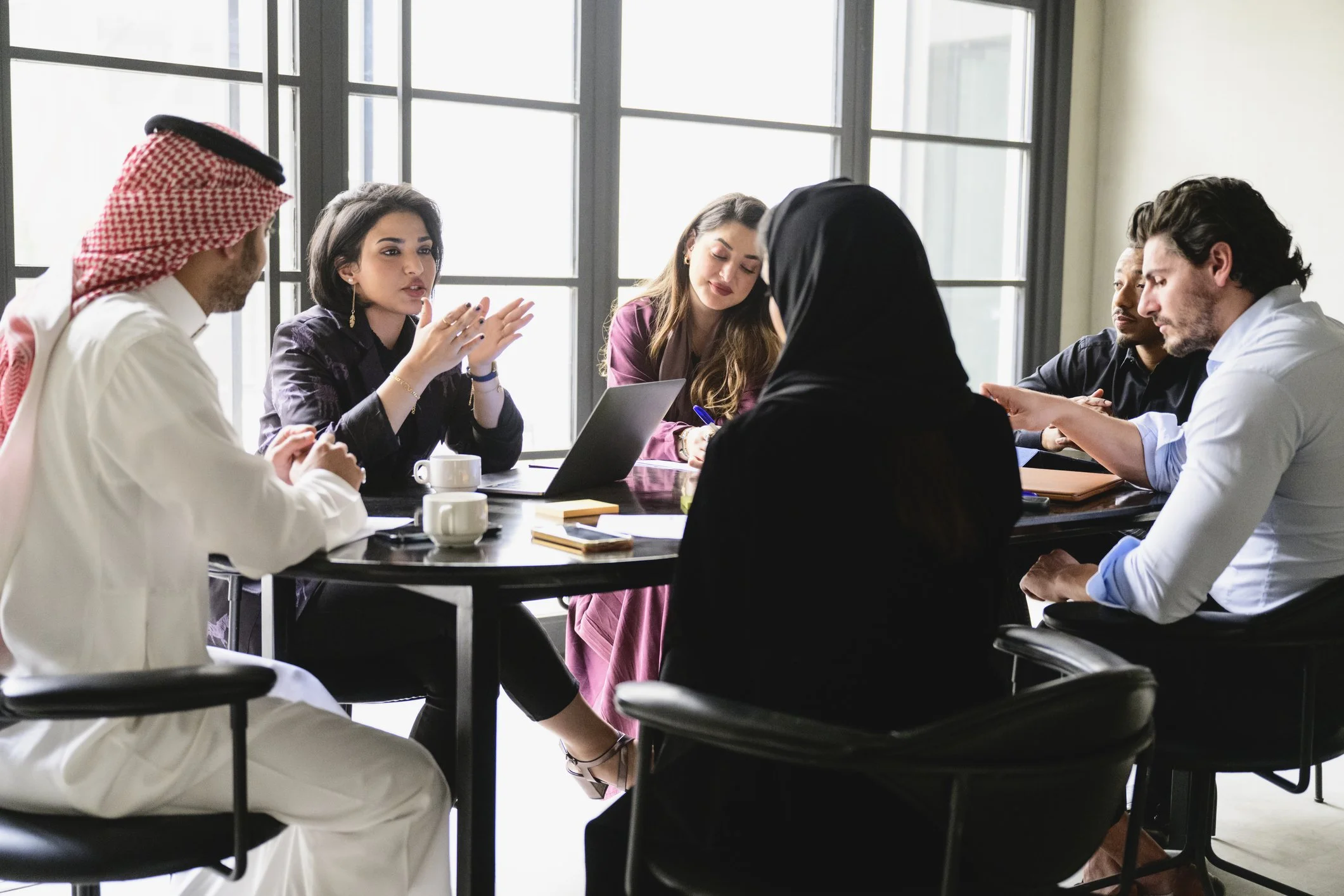 A woman wearing a hijab and a young girl sit together at a kitchen table using a laptop, with the woman holding a credit card and an open book nearby.