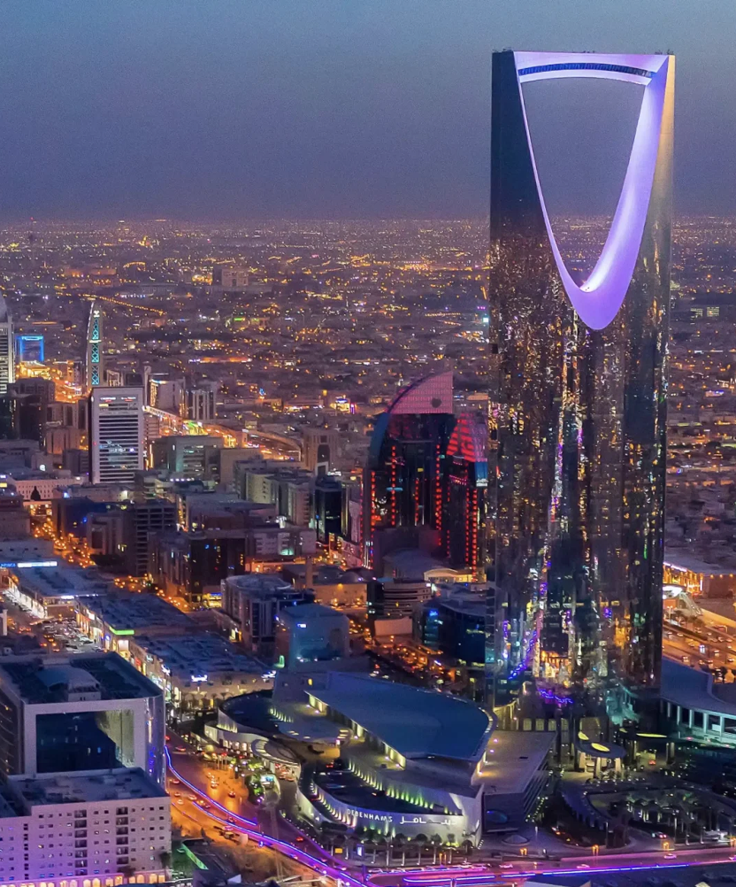Nighttime aerial view of a cityscape with illuminated buildings, including a tall skyscraper with a unique curved top, colorful reflections, and city lights stretching into the distance.