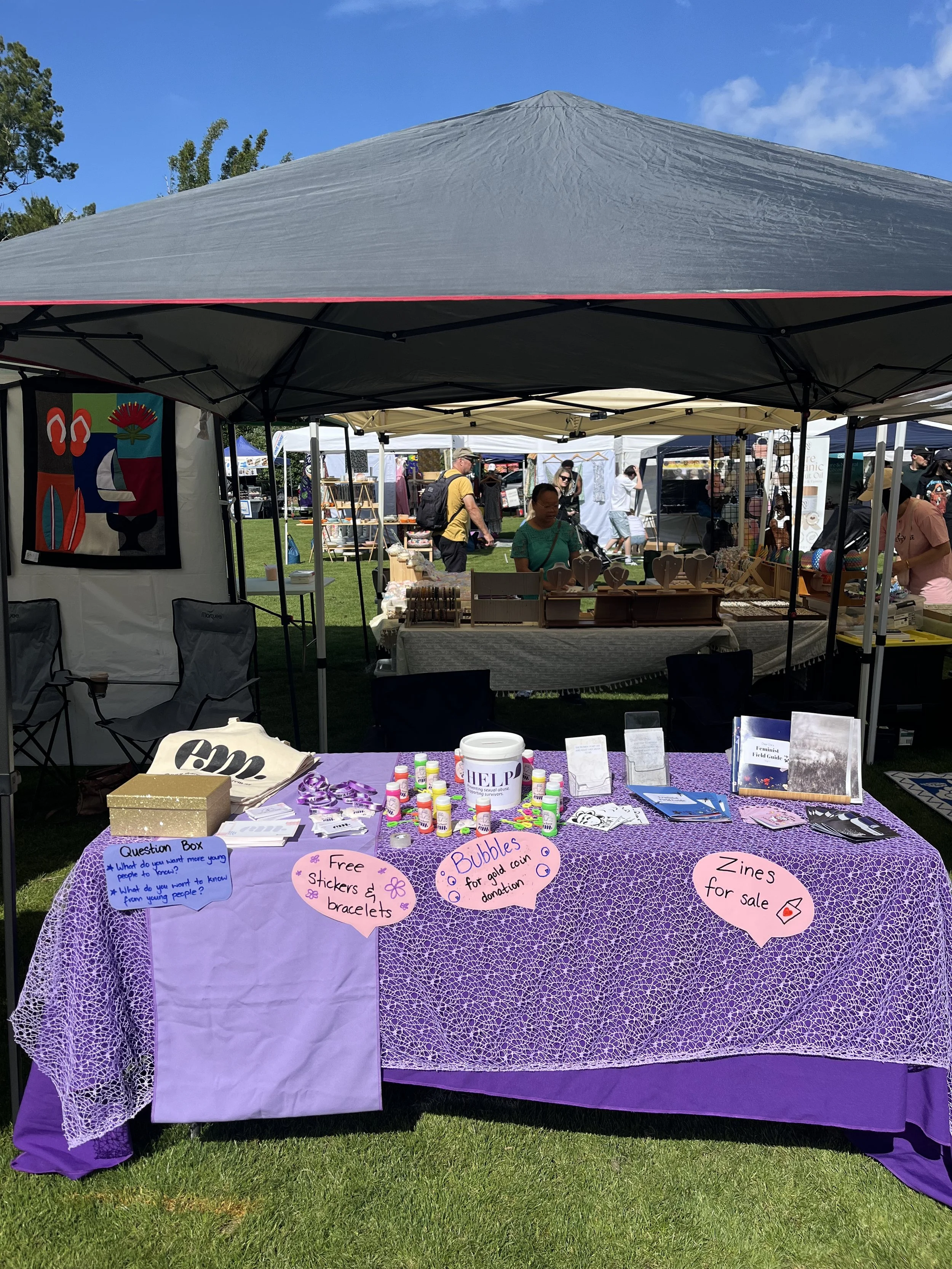 A market stall at an outdoor event, with zines for sale, gold coin donations, and free dear em merchandise