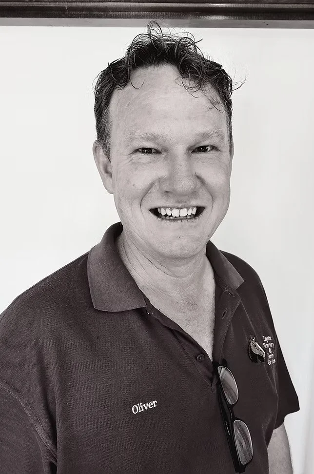A smiling man wearing a polo shirt with the name Oliver and an embroidered logo, standing against a plain background.