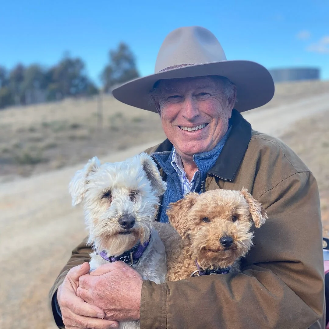 An elderly man smiling and wearing a wide-brimmed hat, holding two small dogs outdoors against a blue sky and distant trees.