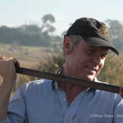 A man wearing a black cap and a light blue button-up shirt is holding a golf club over his shoulder while standing outdoors.