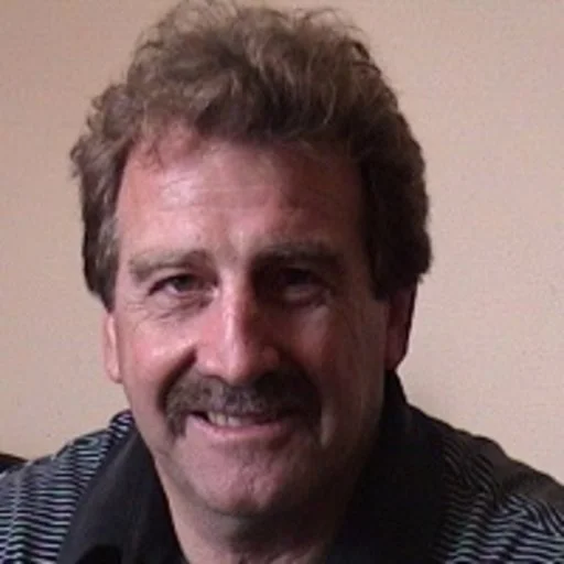 A smiling man with light brown hair, a mustache, wearing a dark striped shirt, sitting indoors with a beige wall background.