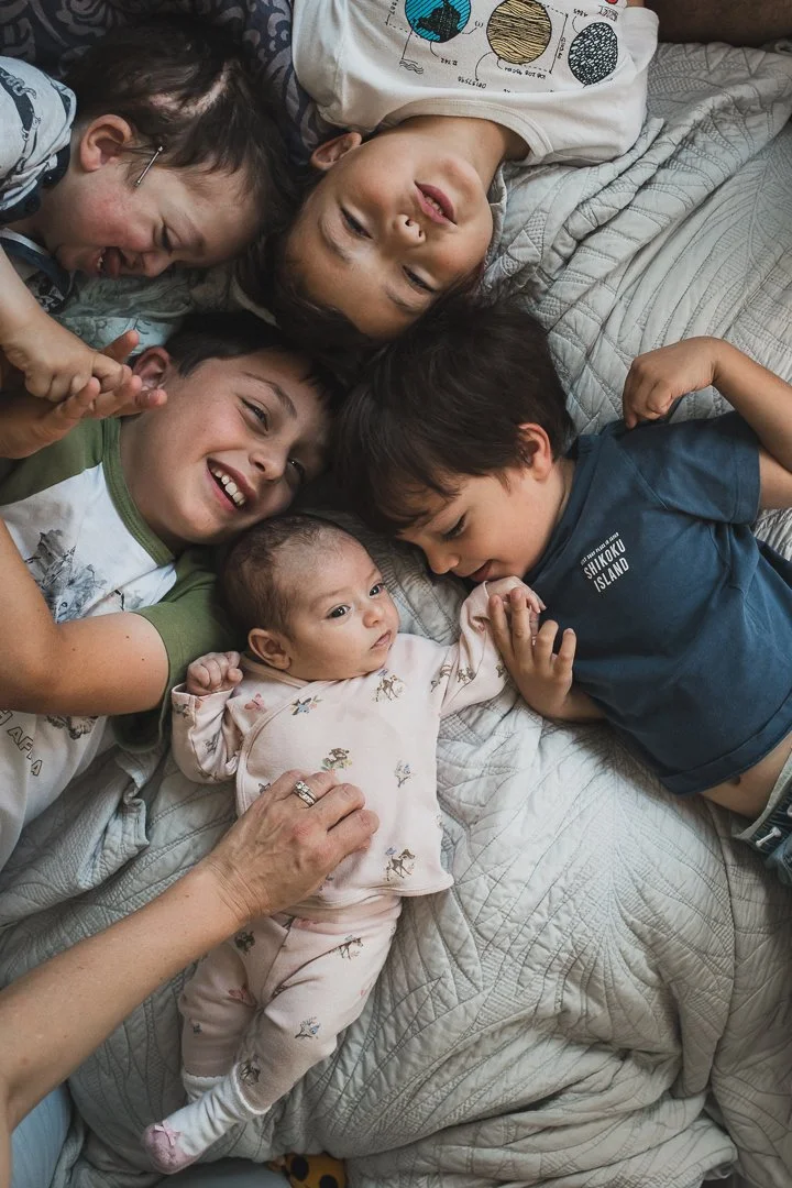 Ein modernes Geschwisterportrait auf dem Bett, von ober fotografiert. 5 Geschwister inklusive ein Neugeborenes Baby liegen Kopf and Kopf.