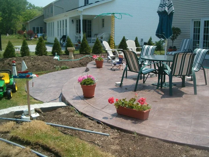 A circular, light-colored, tiered concrete patio stamped to look like stone pavers. A green patio table and chairs are set on it.