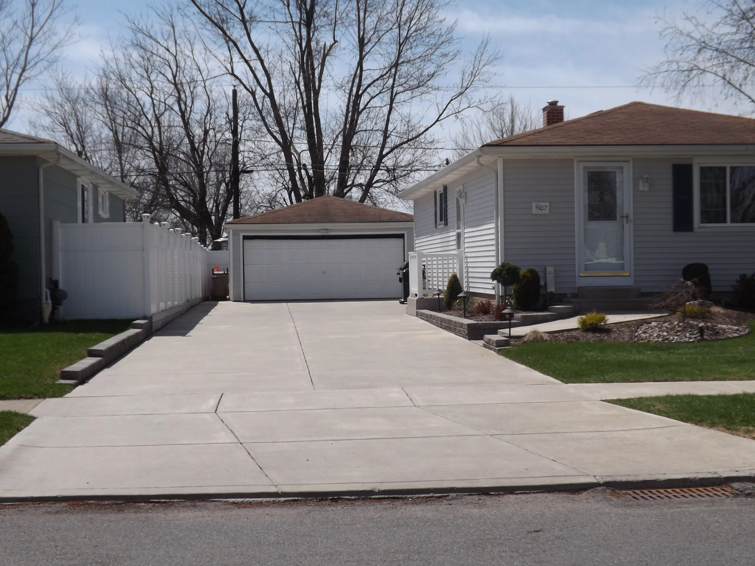 A regularly-colored two-car concrete driveway flanked by a gray house. It leads to a two-car garage.