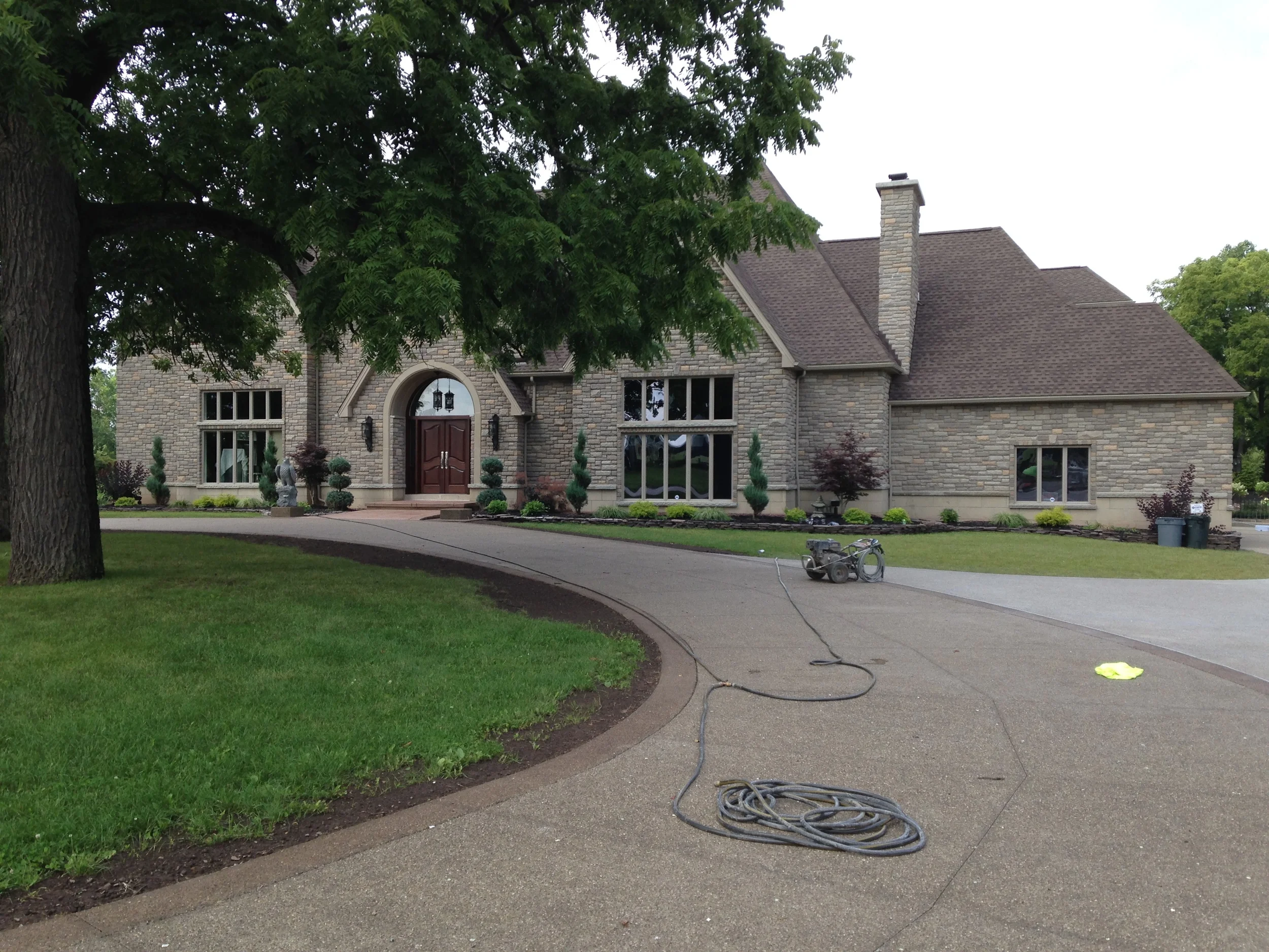 Large concrete wrap-around driveway with dark trim in front of a large stone house. Equipment sits in the driveway.