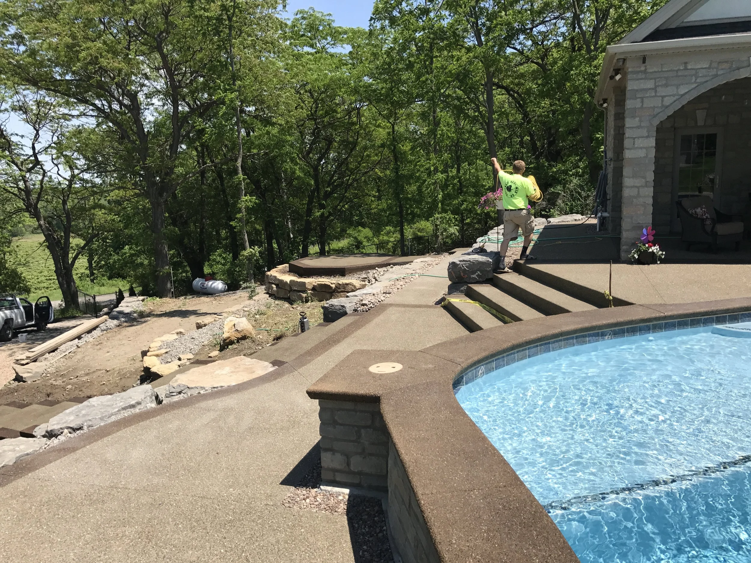 Pool with Waterfall in Buffalo NY. A pool with new a new concrete patio has a waterfall being added beside it. A worker with a bright-green shirt sets up equipment.