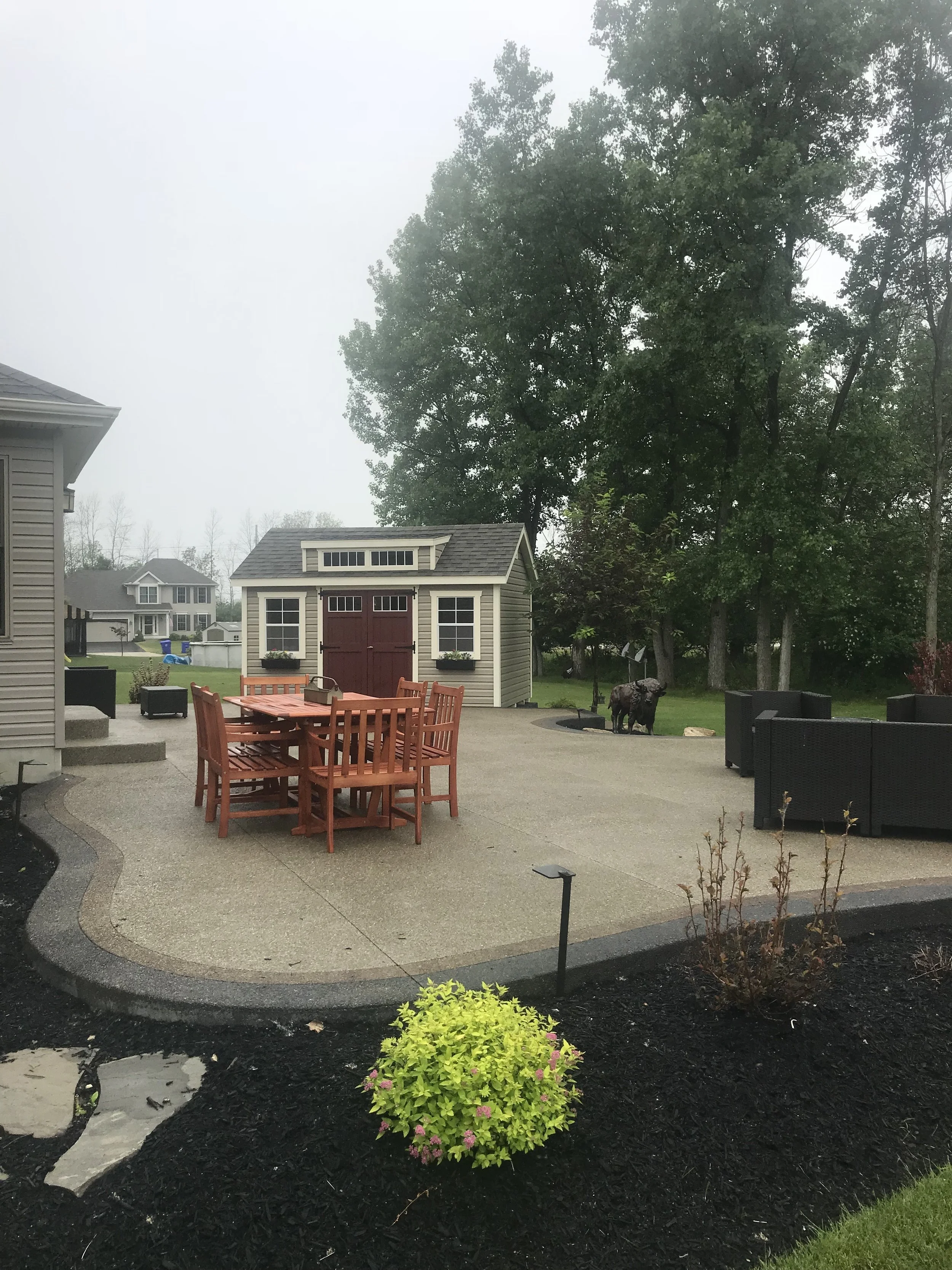 Custom Concrete Patio for your home. A light concrete curved patio. It has patio furniture and a dining set on it, is flanked by gardens and a small gray shed.