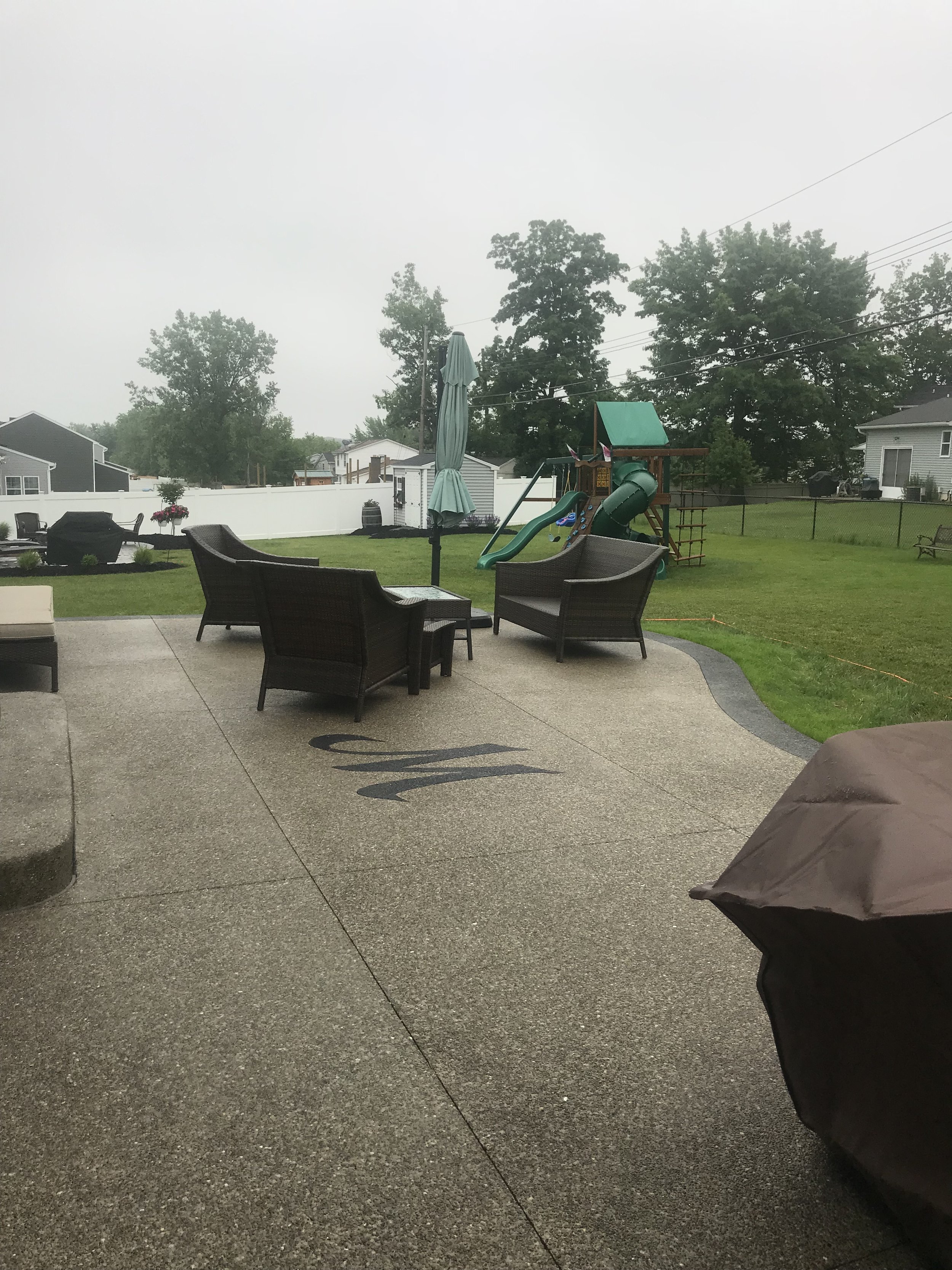 Monogram Patio Backyard. A light colored concrete patio with a monogram M set into it. Wicker furniture and a covered grill are set up on the patio.