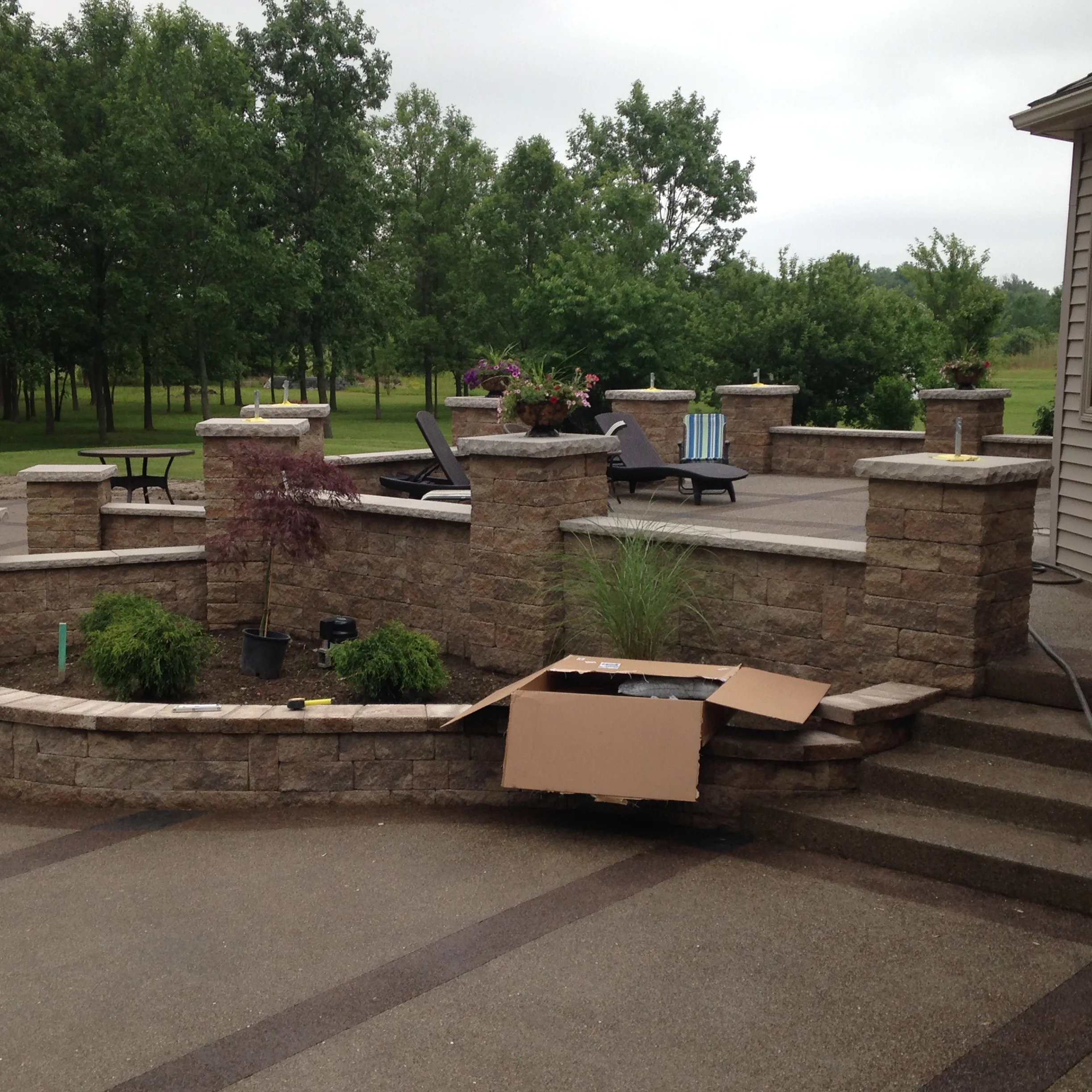 Stone faced concrete retaining wall and steps. Square retaining walls surround a cement patio. Cement steps lead down to a second tier with gardens behind retaining walls.
