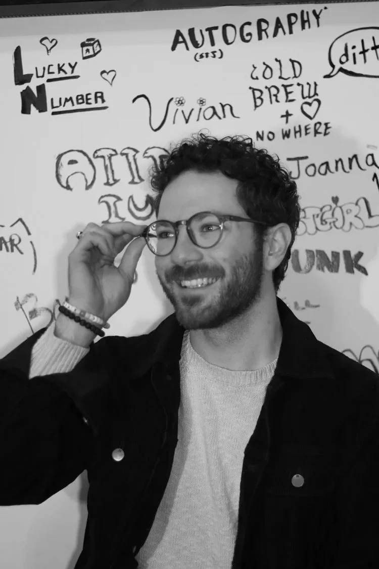 A smiling man with curly hair and glasses adjusting his glasses, standing in front of a wall covered with various handwritten words and phrases in black ink.