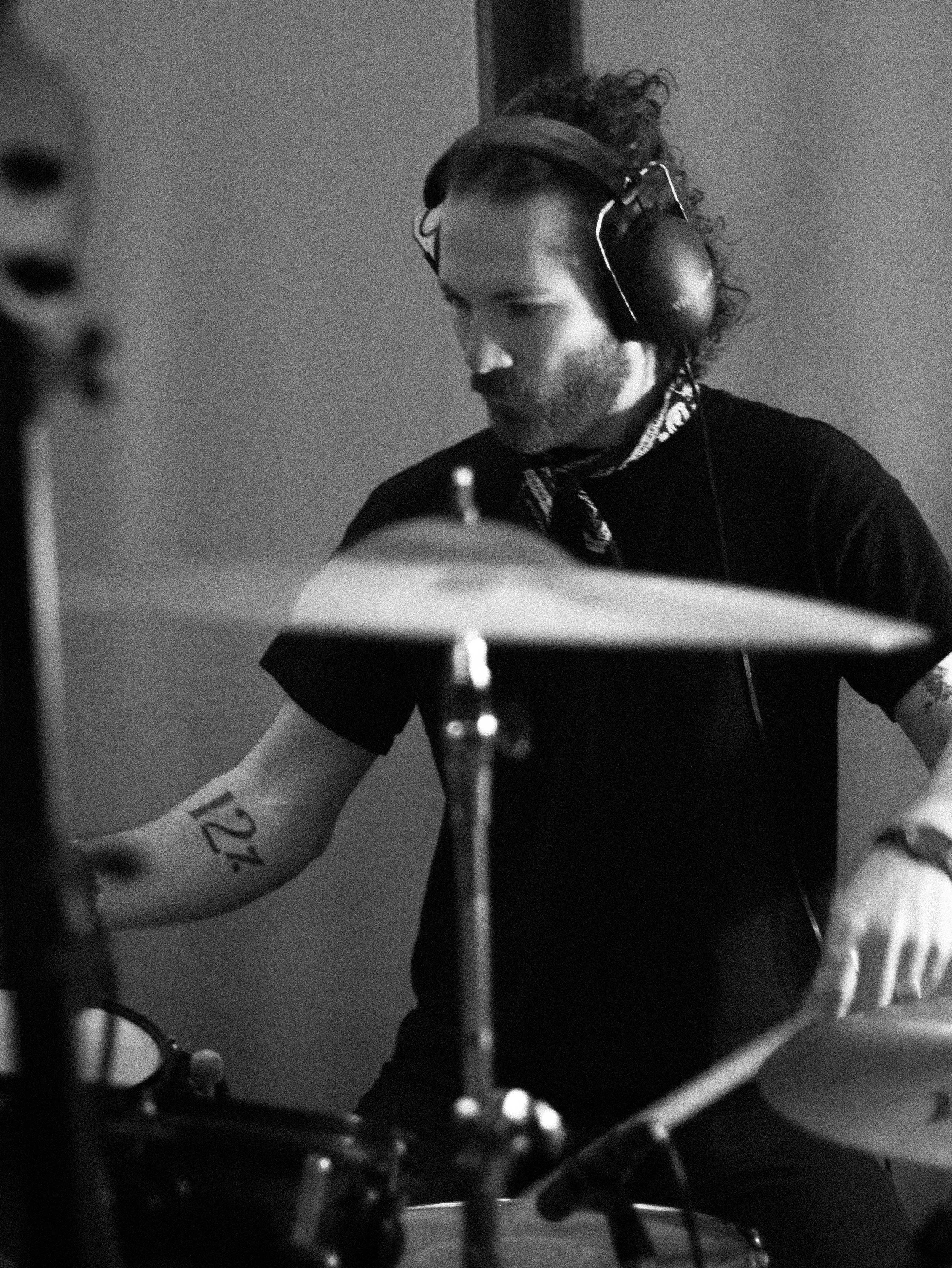 Black and white photo of a man with curly hair and a beard wearing large headphones, looking downwards, with a music stand in front of him and musical notes on his arms.