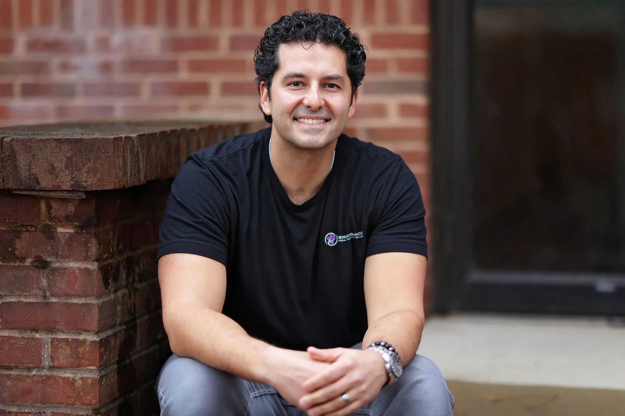 A smiling man with curly dark hair wearing a black V-neck T-shirt, standing against a plain light gray background.