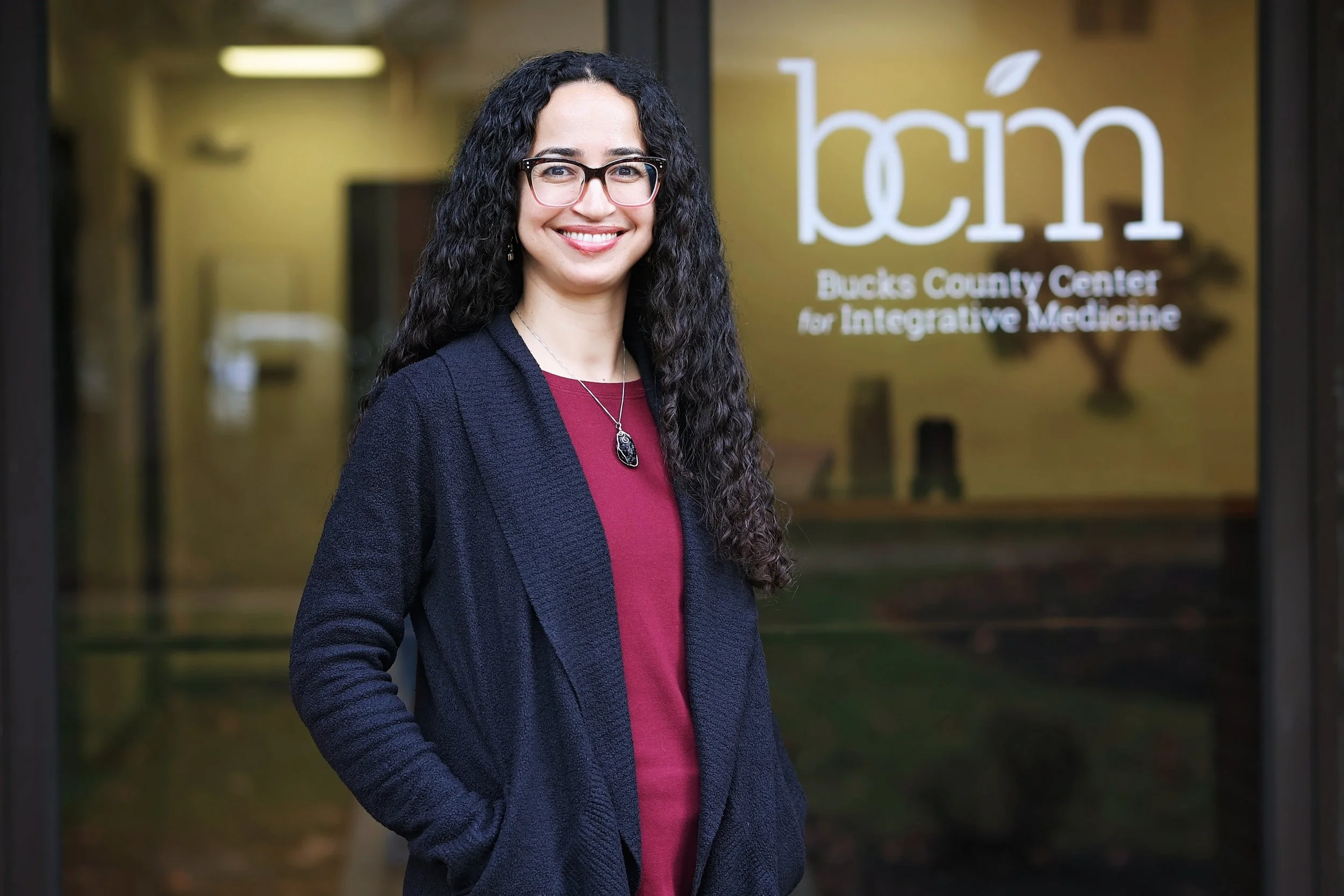 A woman with curly dark hair, glasses, and a necklace, smiling while standing against a brick wall.
