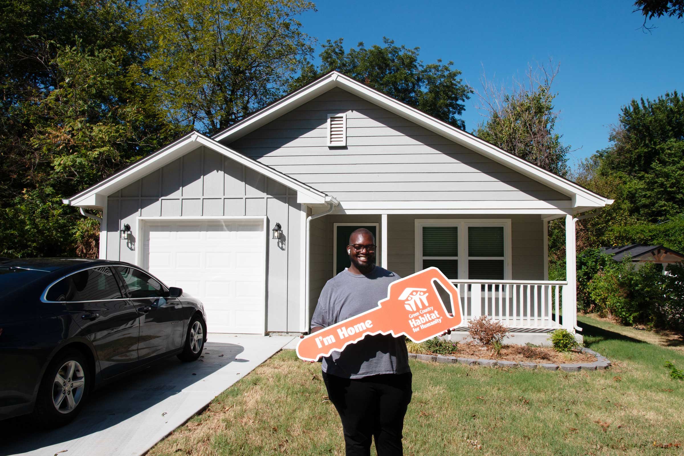 AFRICAN AMERICAN MAN HOLDING AN OVERSIZED KEY AND SMILING IN FRONT OF HIS NEW HOME