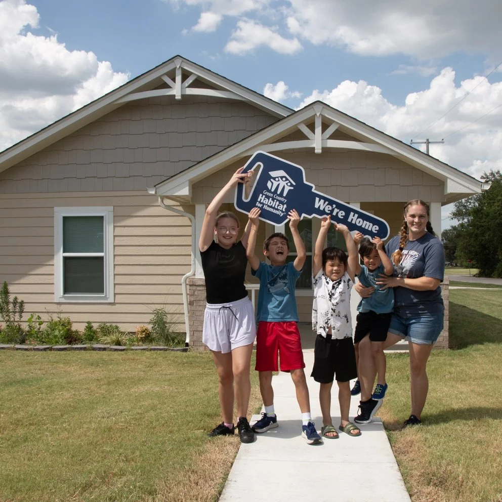 a young mother and her four children stand in front of their new home halding an oversized key with the Green Country Habitat for Humanity logo and the words "We're Home"