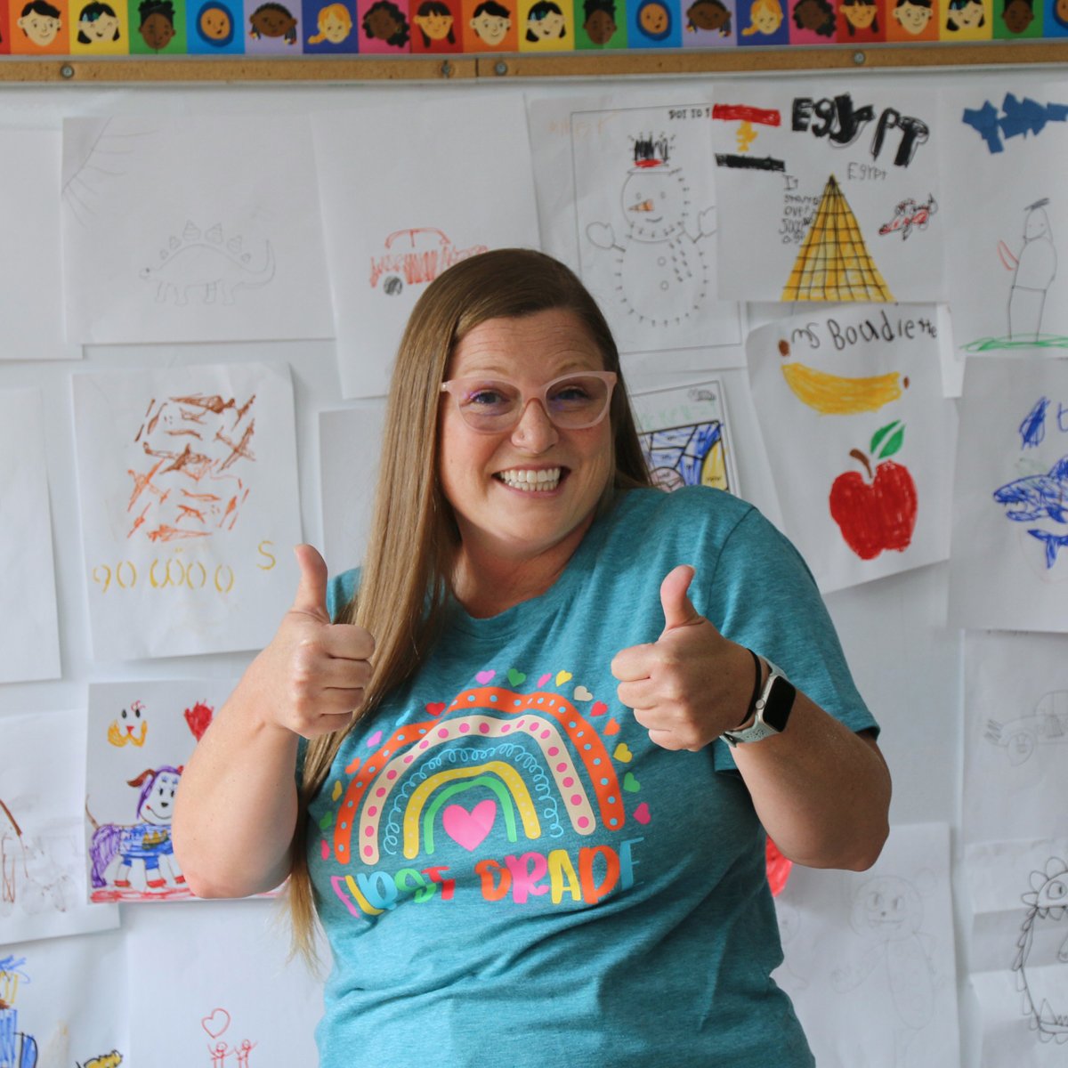 a teacher gives the thumbs up standing in front of a bulletin board full of student drawings