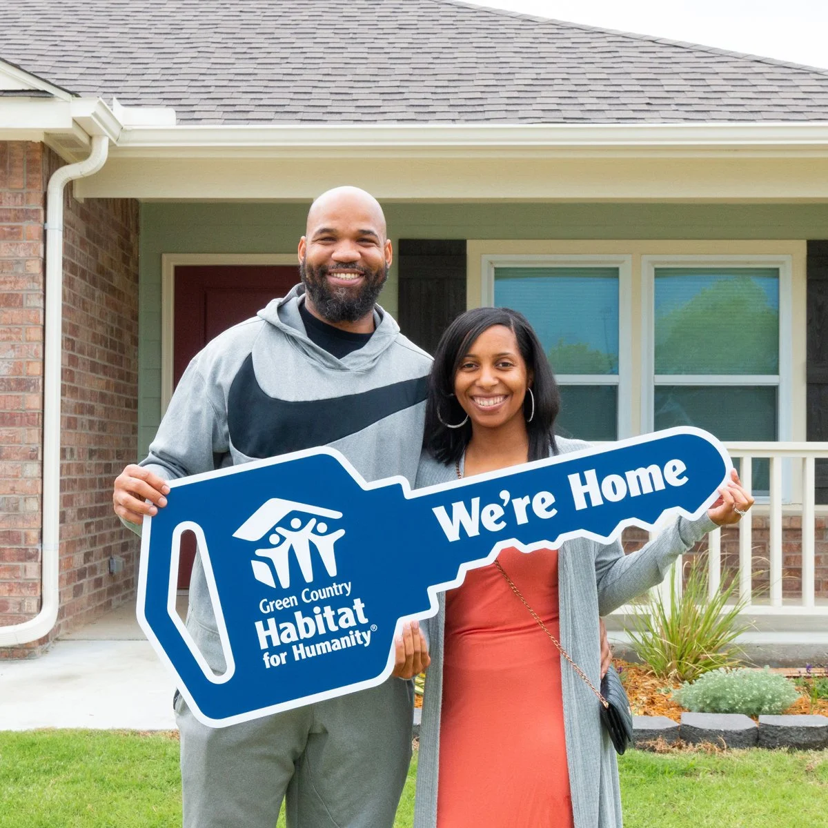 An african american couple stands in front of their new home holding an oversized key with the Green Country Habitat for Humanity logo and the words "We're Home"