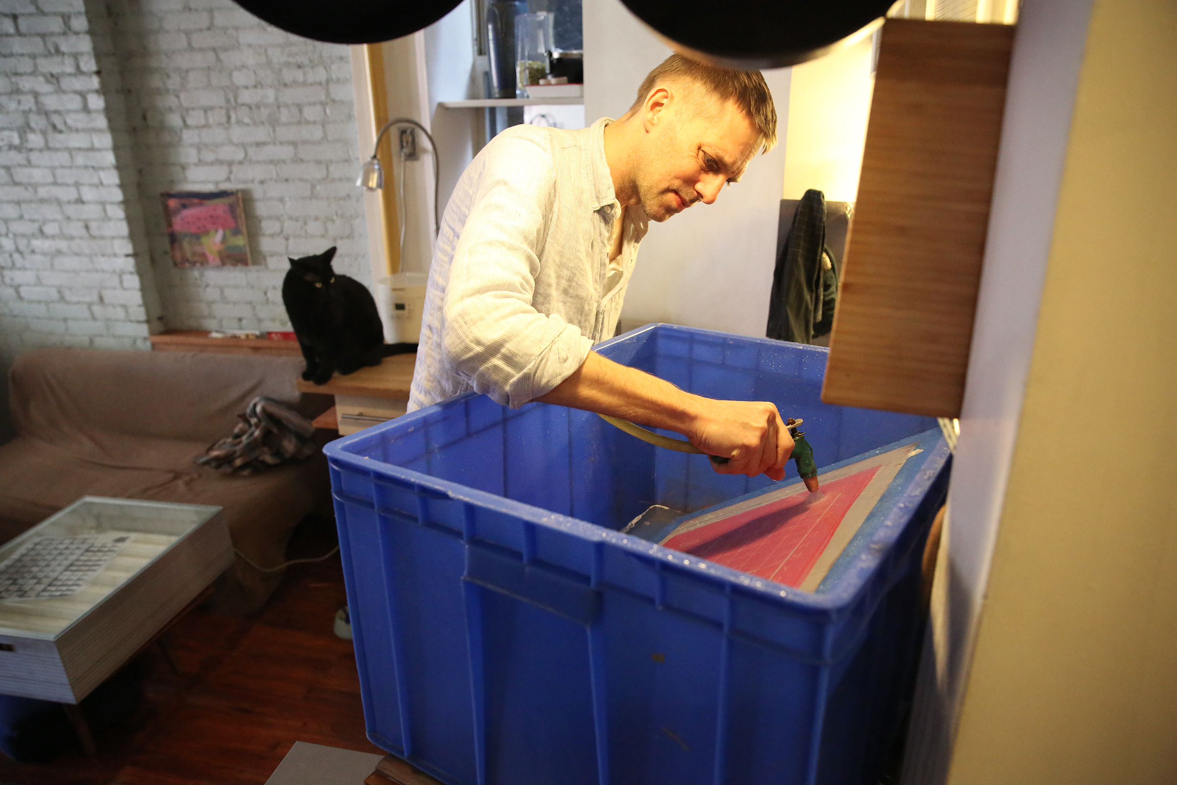 Anselm washes ink off of the Japanese language typography screen. The kitchen is very organized with everything stored or hung in place. Pans and a cutting board can be seen around the sink.