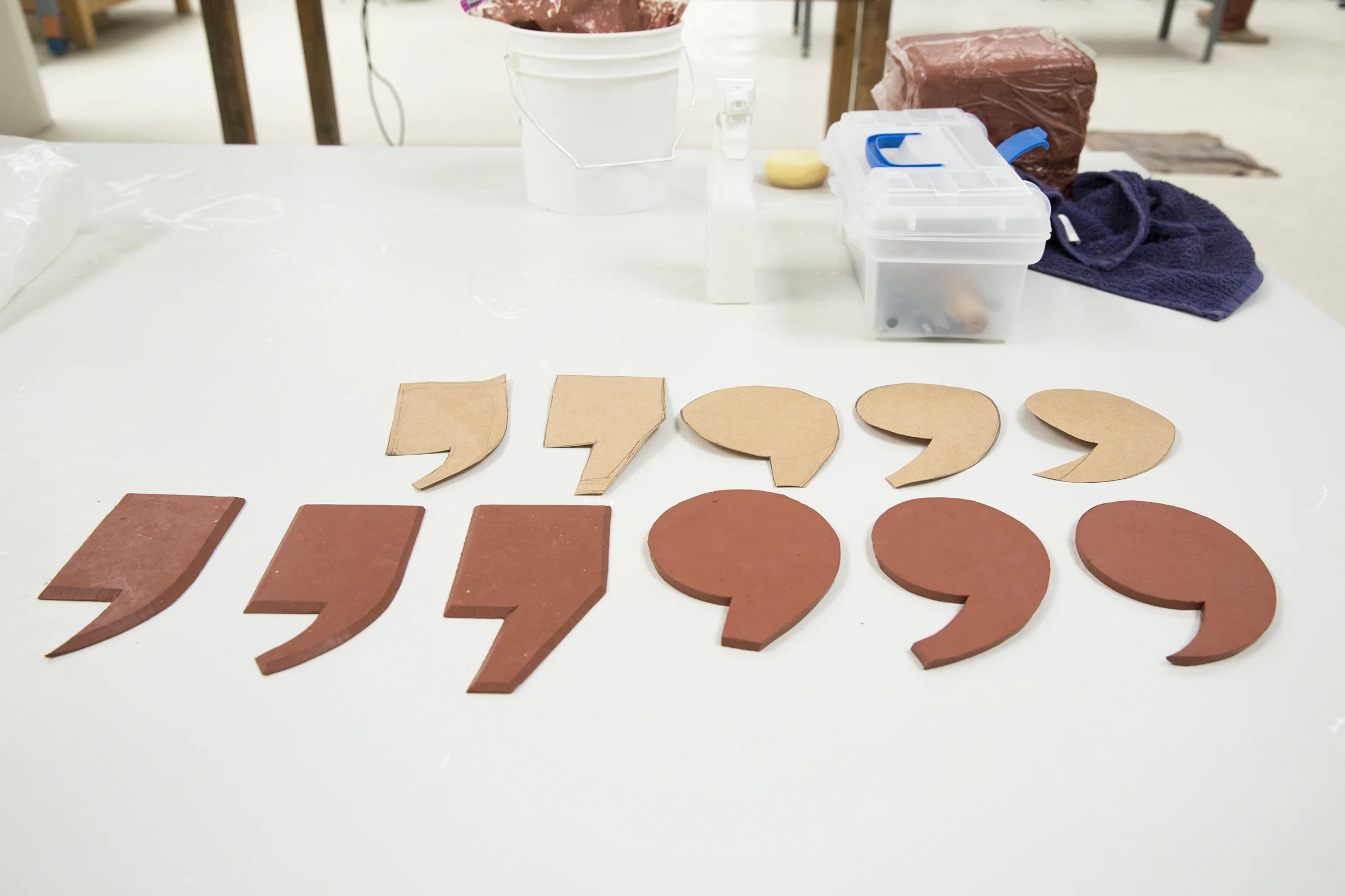  Paper patterns that Karolina used to trace commas and quotation marks sit above the clay pieces. These will become plates that will be glazed solid black. Sculpting tools and stoneware clay are on the work table. 