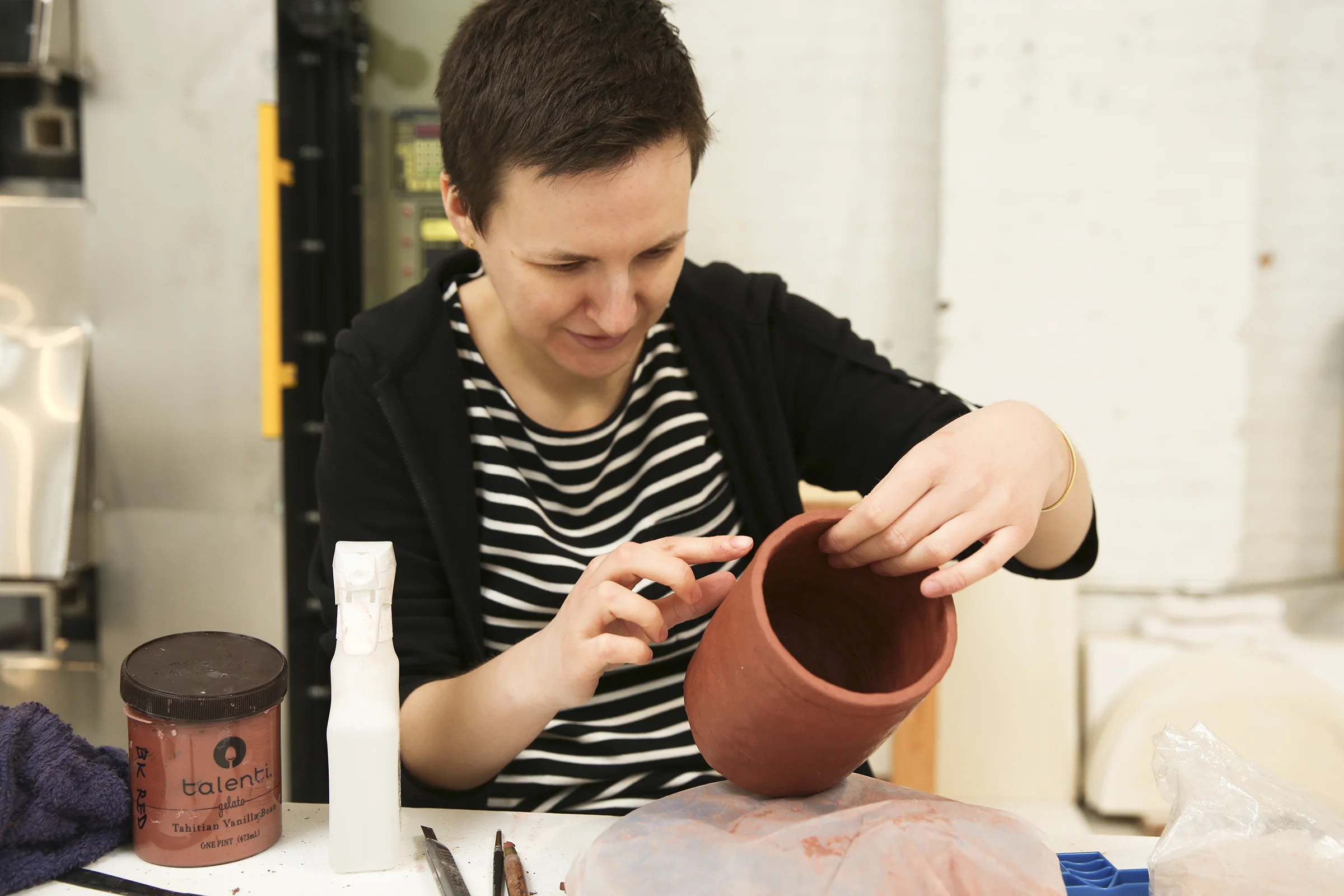  Karolina uses her finger to smooth slip onto the wet clay mug body. A gelato container full of slip (a mixture of clay and water) and a spray bottle of water to help join and bind sections of clay and evenly smooth over sections.  