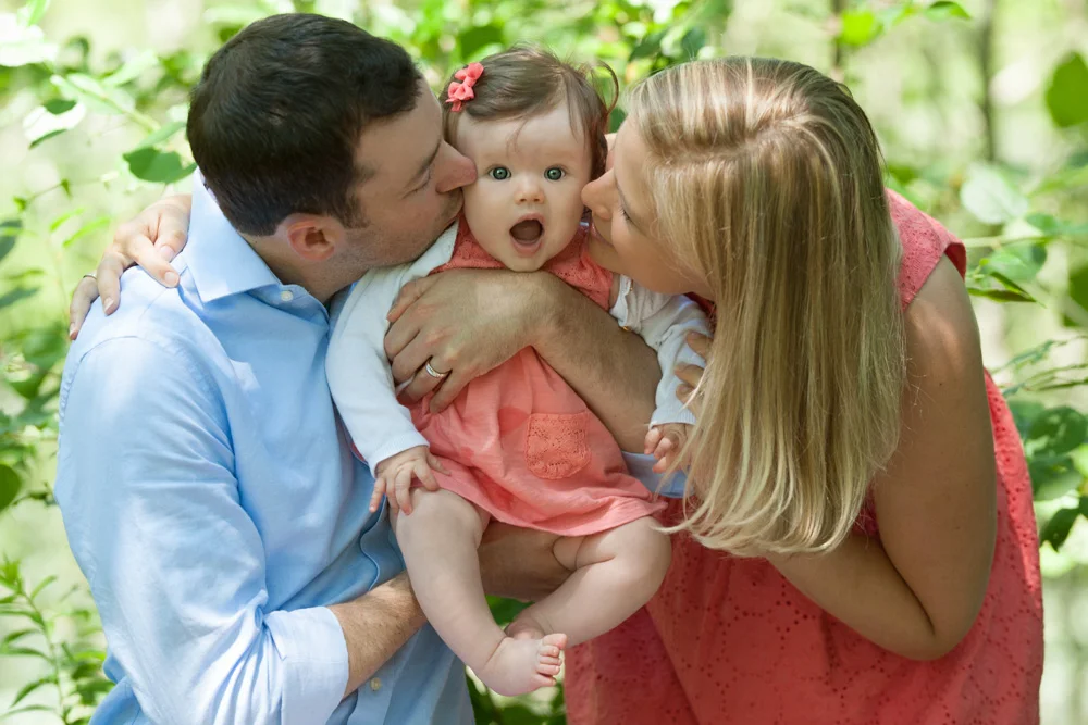 Mother and Father kissing adorable baby looking at camera in sur