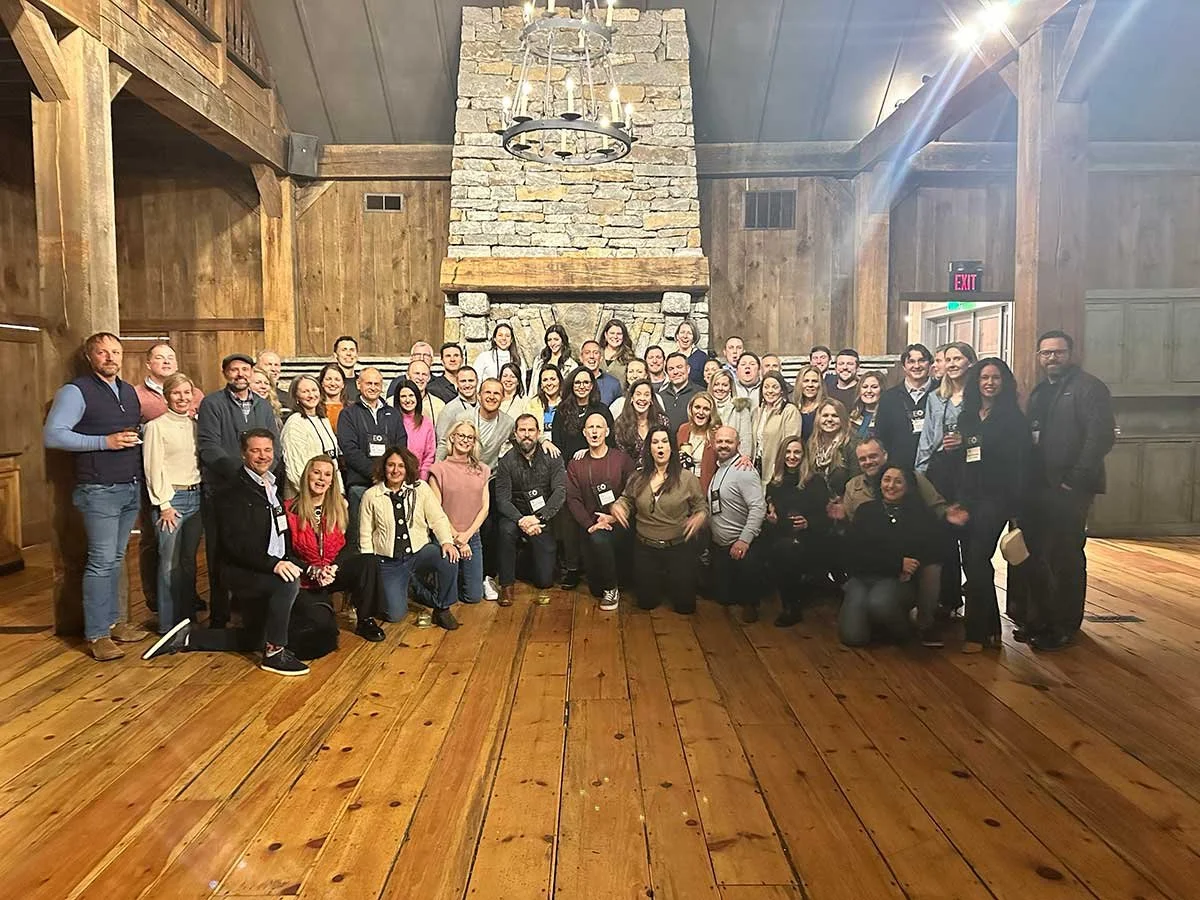 Group of people gathered in a rustic wooden room with a large stone fireplace and wood-paneled walls.