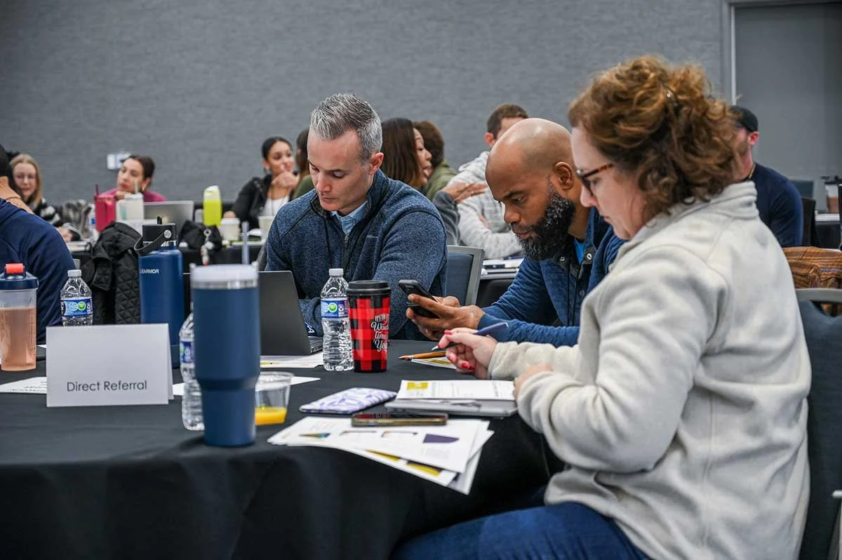 Group of people seated at a conference table, some looking at electronic devices, with papers, water bottles, and cups in front of them, in a professional setting.
