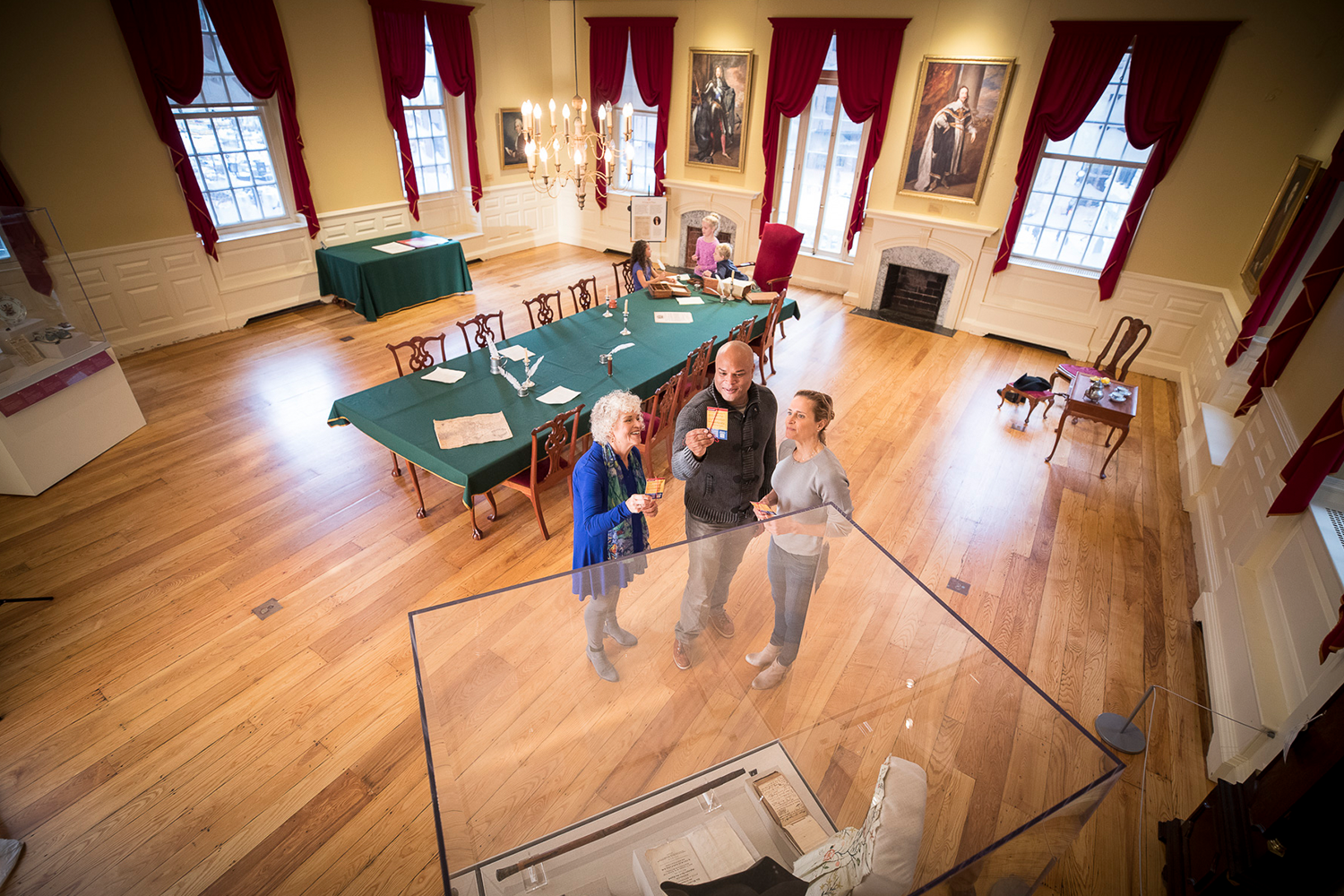 Three adults examining artifacts in a display case while children sit at the Council Chamber table.