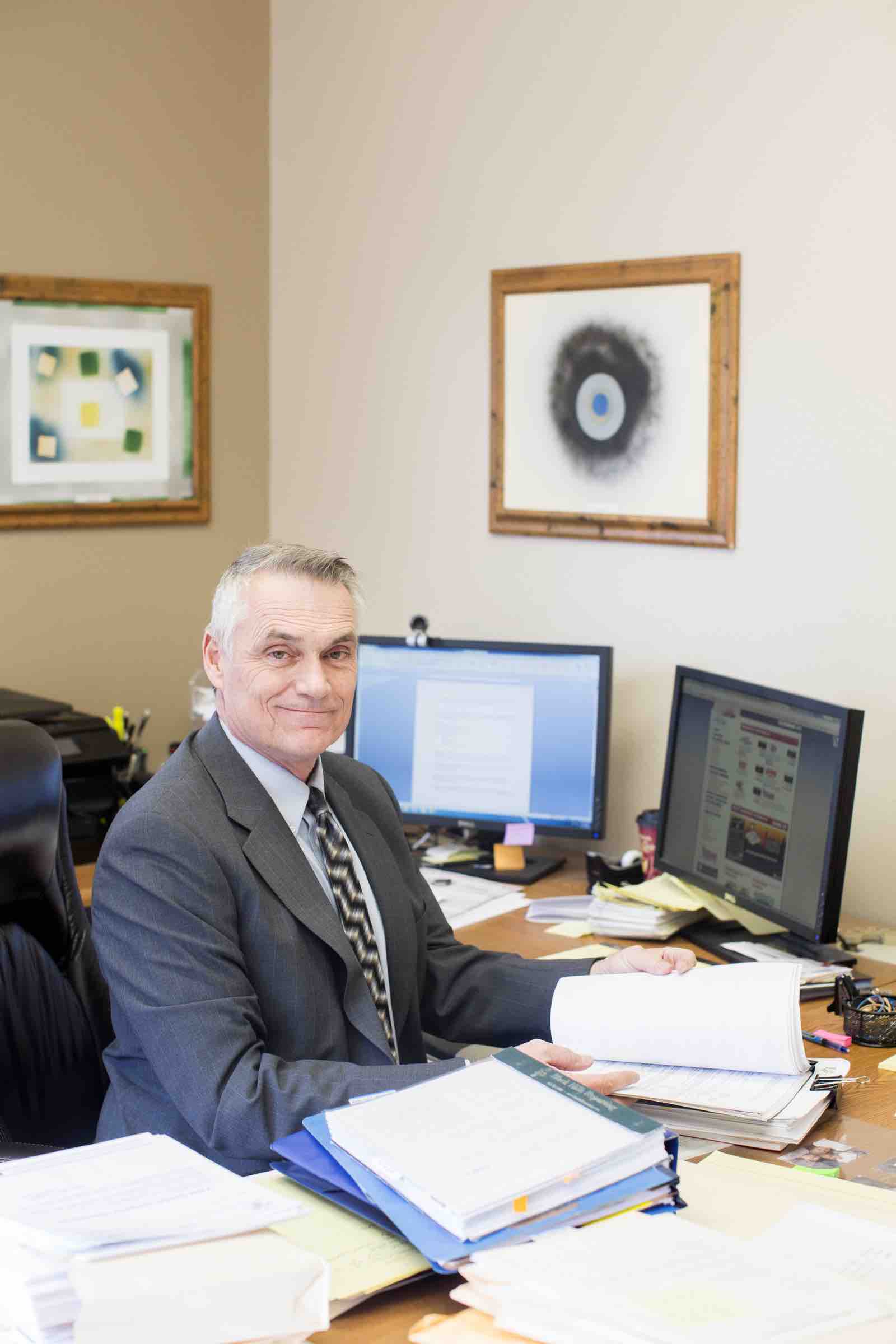 George Nelson at his desk with dual monitors and wall hanging photos