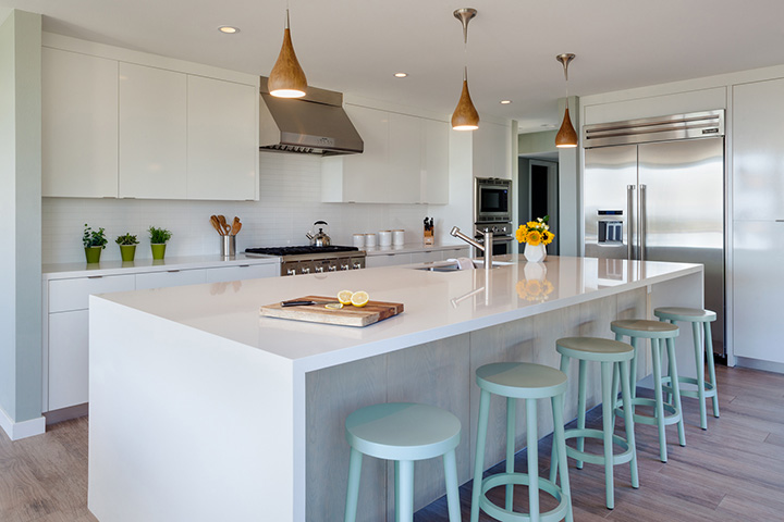 With a white kitchen, light mint stools play a big role in creating a cozy atmosphere. Waterfall edges on the countertops designate the kitchen area from the dining room space.