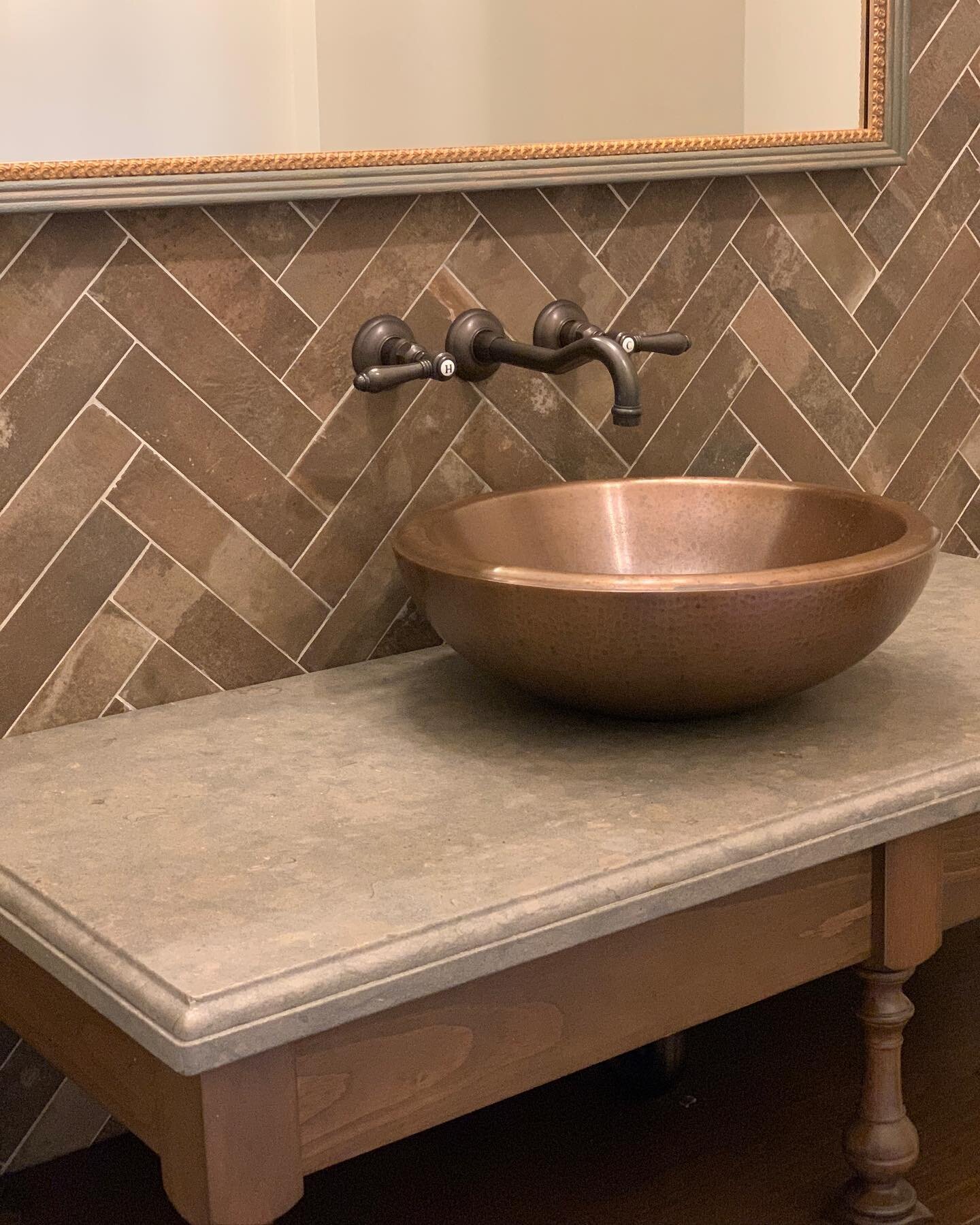What I love about this Powder Room we designed are the layers of materials and textures. A copper sink sits on a stone counter, grounded by an oak console. Behind the bronze faucet and framed mirror the brick-like tile in the herringbone pattern prov