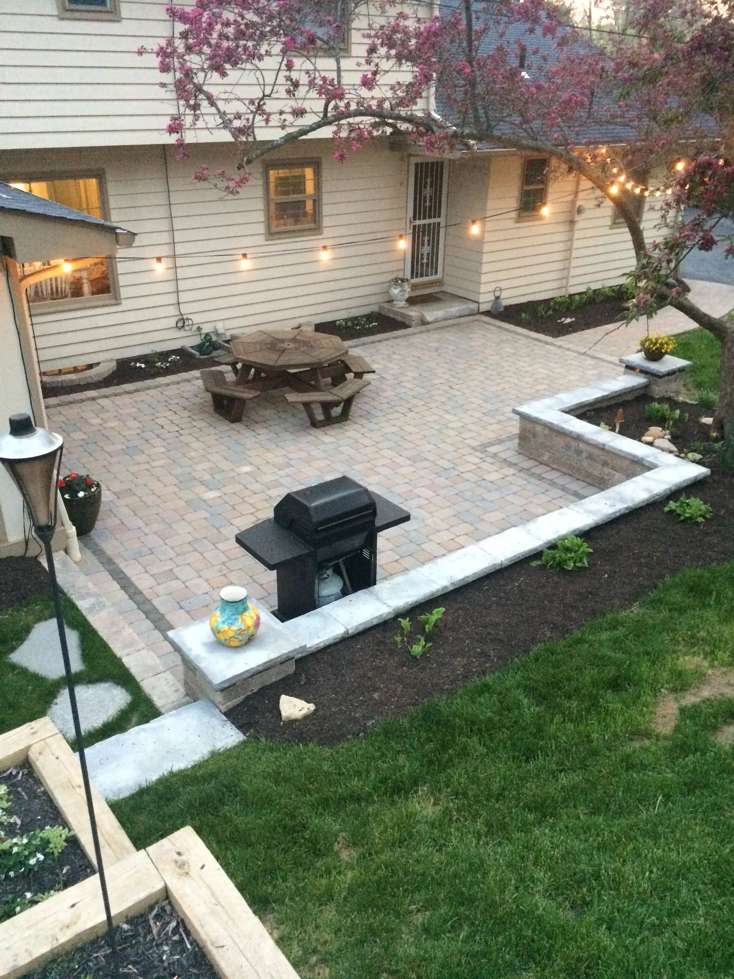 Backyard patio with brick flooring, wooden picnic table, barbecue grill, garden with grass and plants, stone wall planters, string lights, and blooming trees.