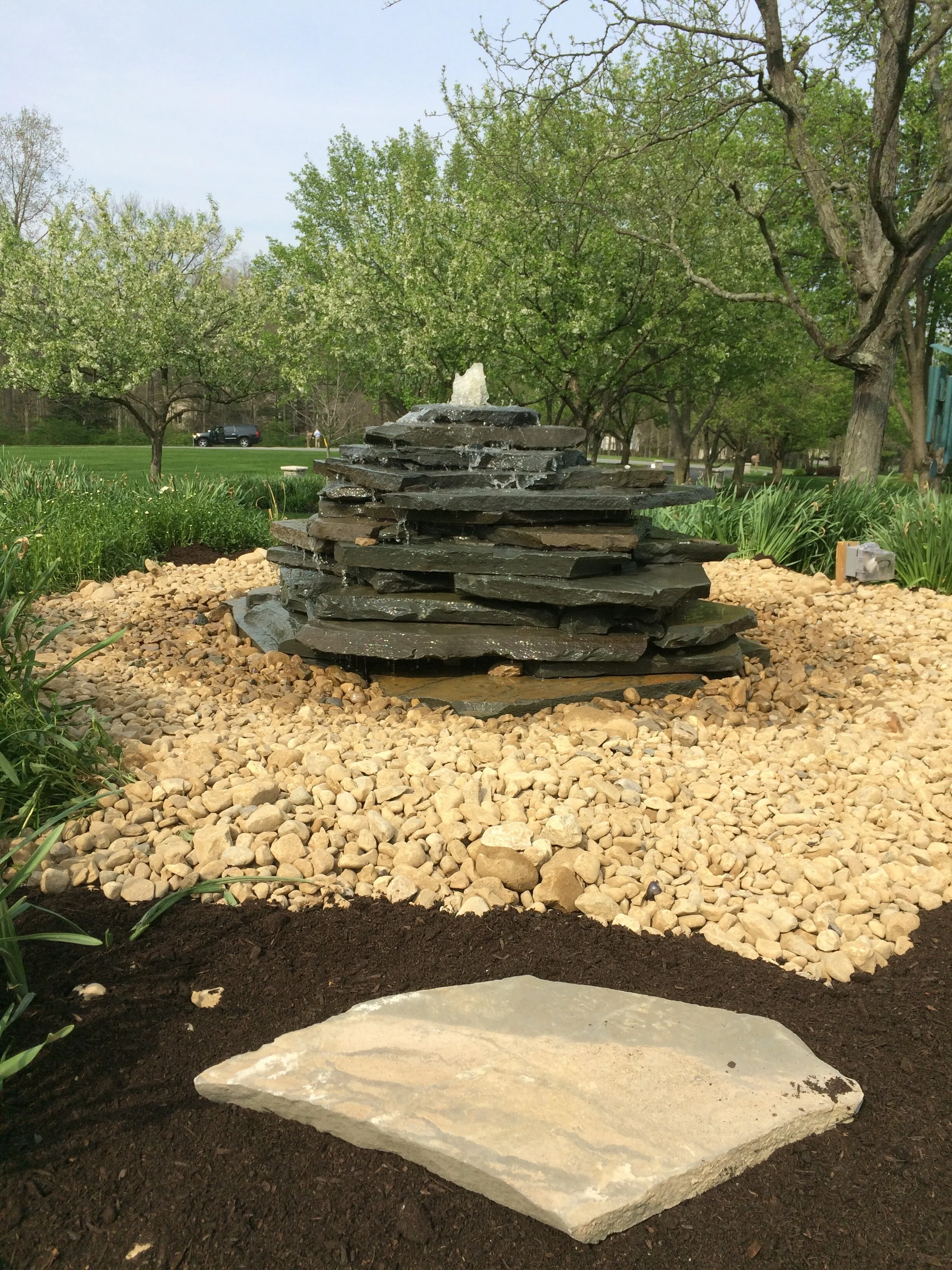 Stacked stone fountain surrounded by pebbles and grass in a park setting with trees in the background.