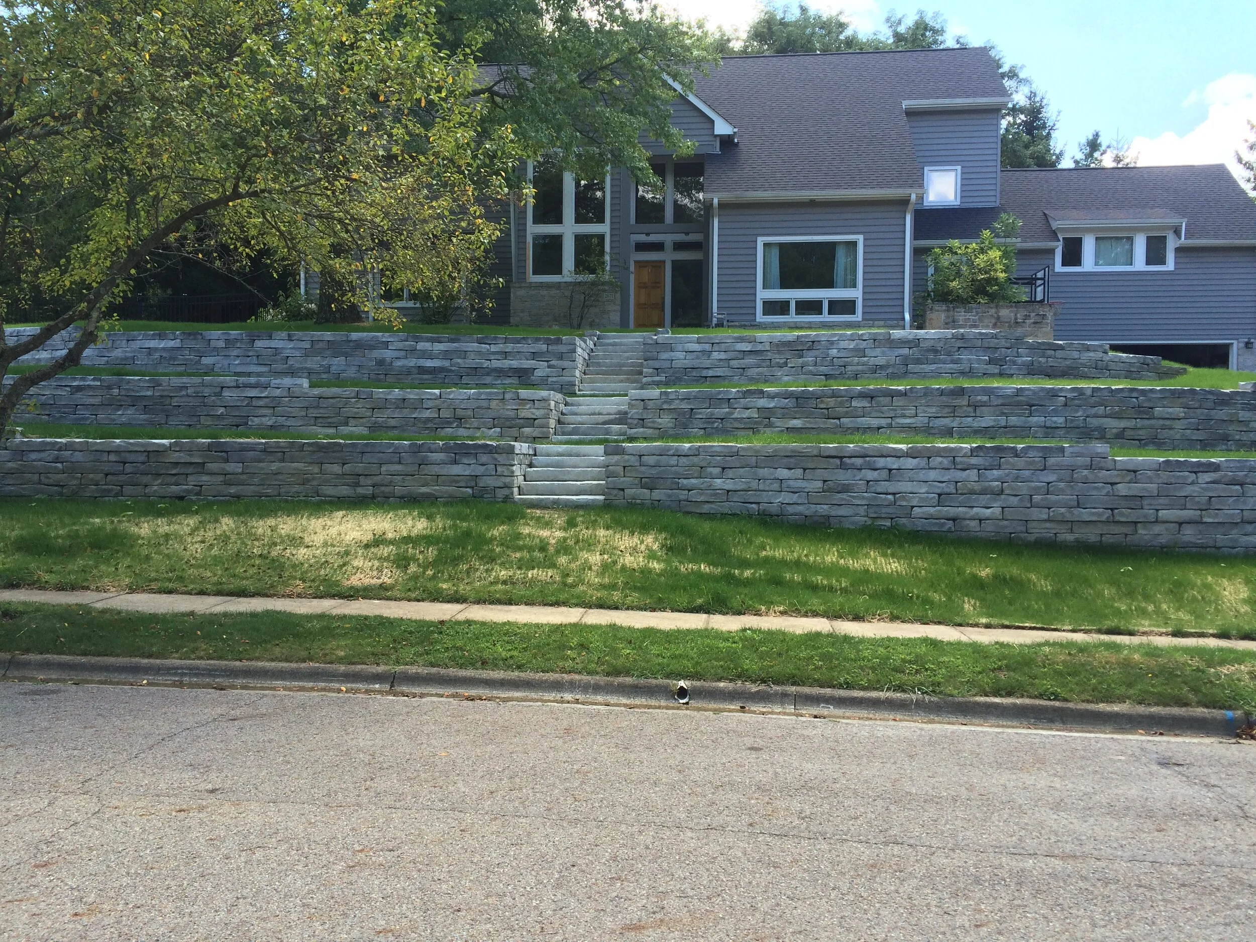 Modern house with gray siding and a large front glass door, surrounded by multiple levels of stone retaining walls and a well-maintained lawn, with a tree on the left.