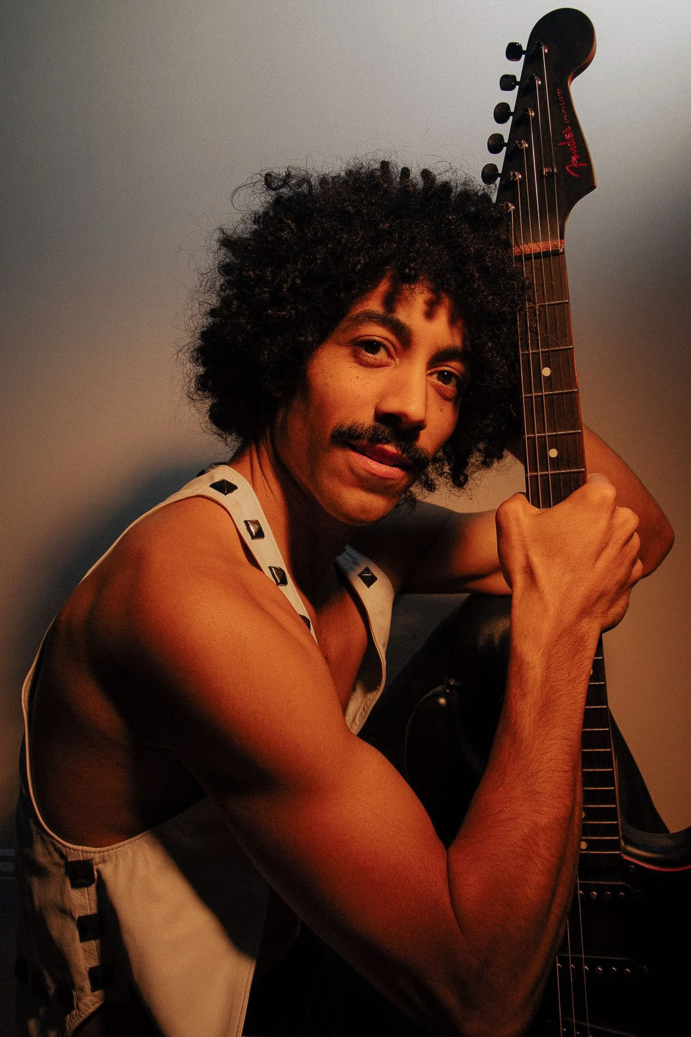 Joe Brown: A man with curly hair, a mustache, and a slight beard holding a black electric guitar, looking into the camera. He is wearing a sleeveless top with metal embellishments on the straps. The background is plain and muted.