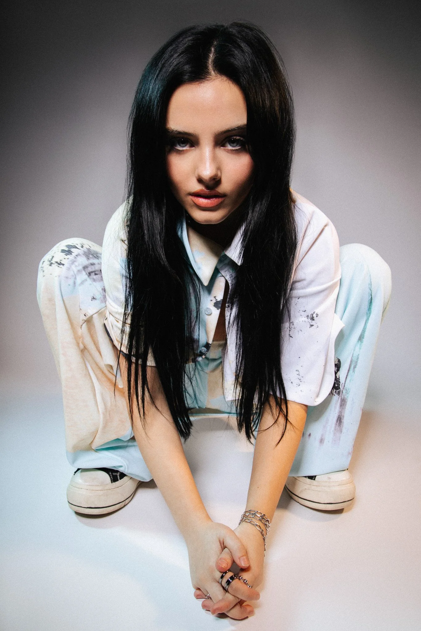 A young woman with long black hair and blue eyes is crouching against a plain, gradient gray background. She is wearing a white, slightly oversized shirt with watercolor-style splashes of color, and light-colored pants. She looks directly at the camera with a serious expression, and her right hand is holding the left hand of the viewer, which is extended towards her. She accessorizes with multiple bracelets and rings.