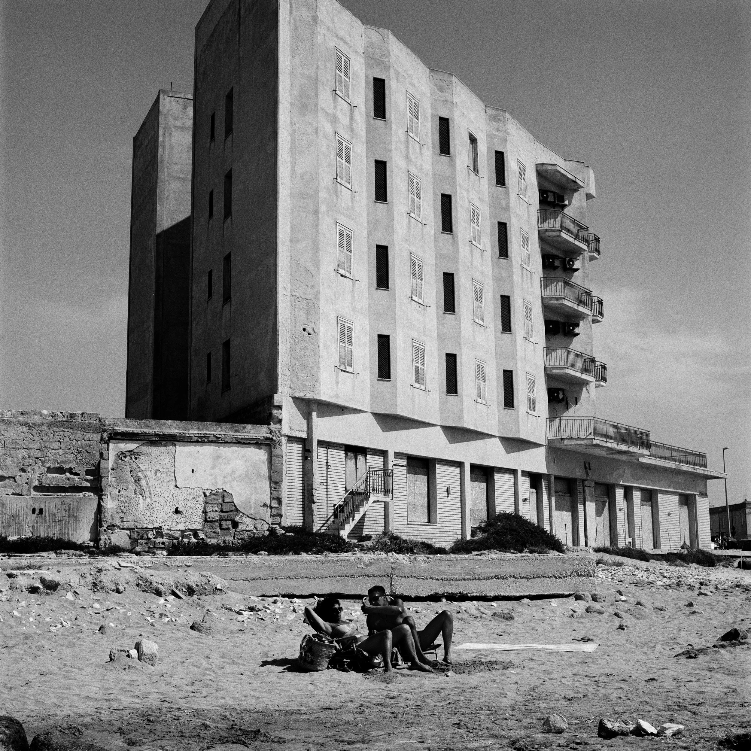  A couple chats along while sun-bathing in front of a foreclosed hotel in Trapani periphery. 