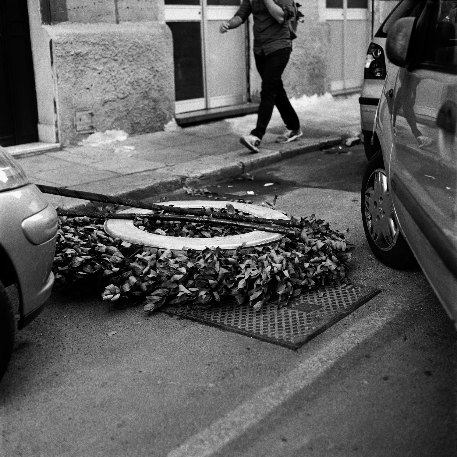 A large funeral wreath lies dumped face-down in the gutter between two parked cars, leaves dried and curling, a broken wooden pole laid across it, half of it resting on a storm drain; in the upper third a pedestrian walks past, cropped so only legs, one arm and a shoulder bag show