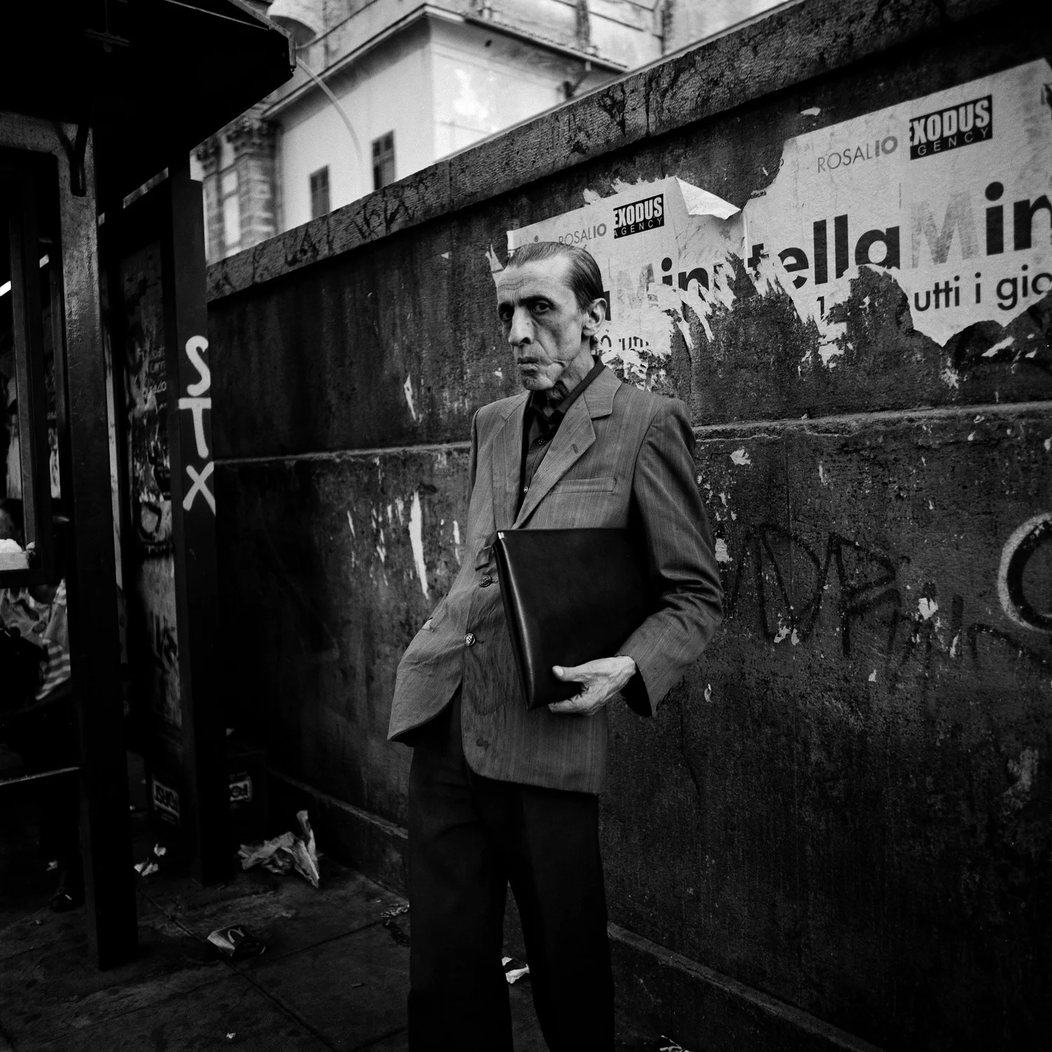 A gaunt hollow-cheeked older man in a rumpled wide-lapelled grey suit stands pressed against a filthy concrete wall clutching a black document folder to his chest, staring flat past the lens; the wall is scabbed with torn advertising, stickers and graffiti, the rusted frame of a bus shelter cutting the left edge