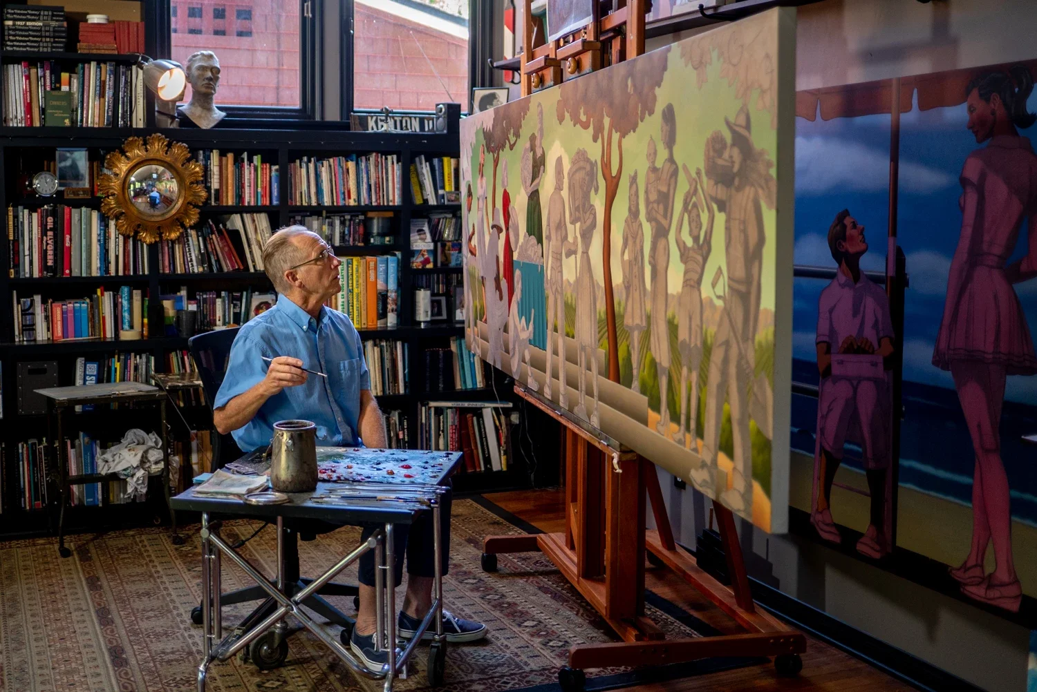 An elderly man sitting in front of a large canvas painting in an art studio filled with books and art supplies, with paintings of people on the canvas and a bookshelf behind him.