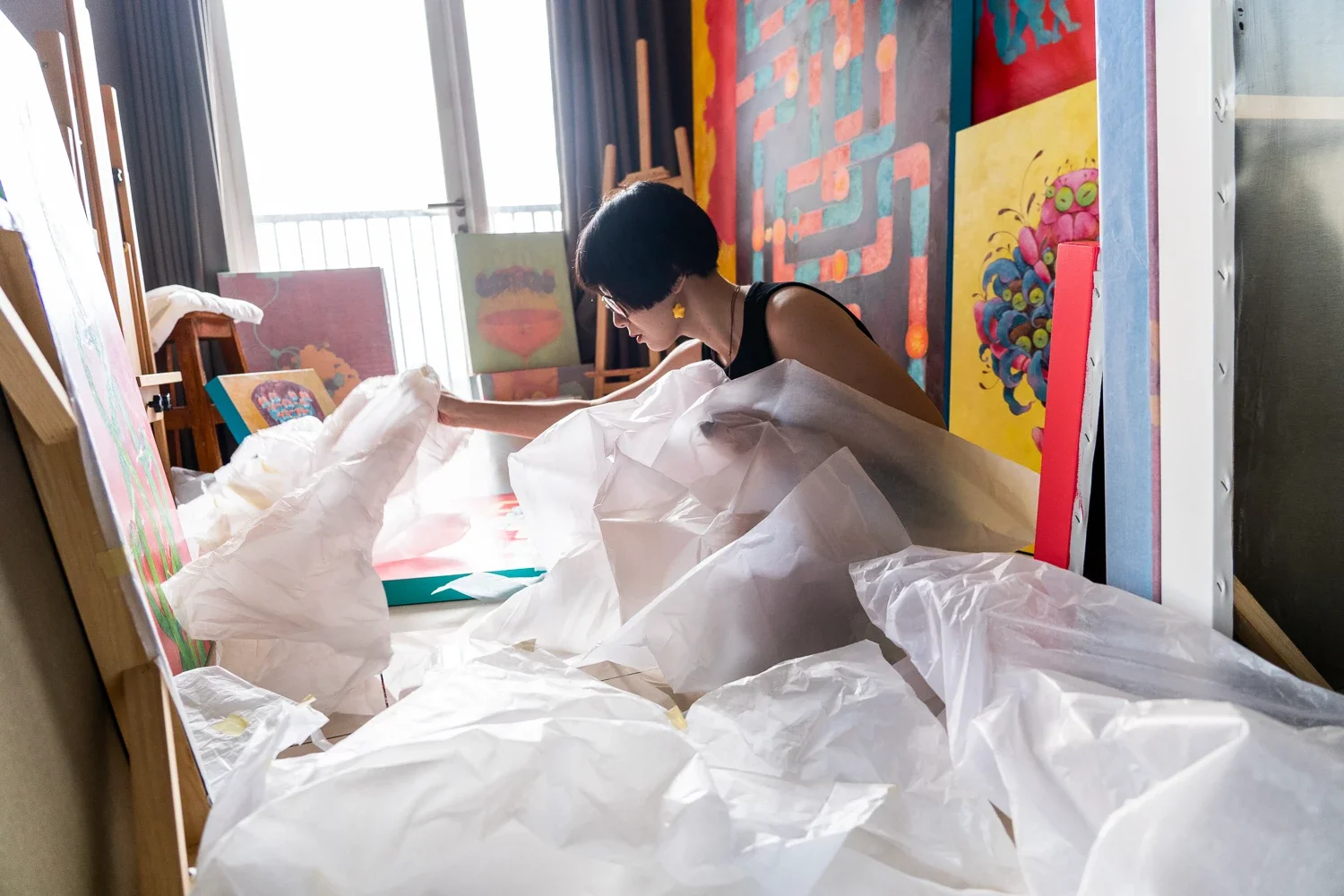An artist arranges colorful paintings in a studio filled with natural light, abstract artworks on the walls and easels.