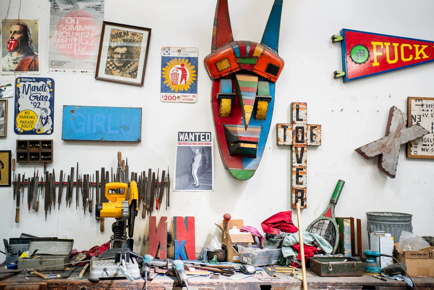 miscellaneous colorful metal artworks and paper clippings on a white wall on top of a wooden workbench with tools and metal pieces